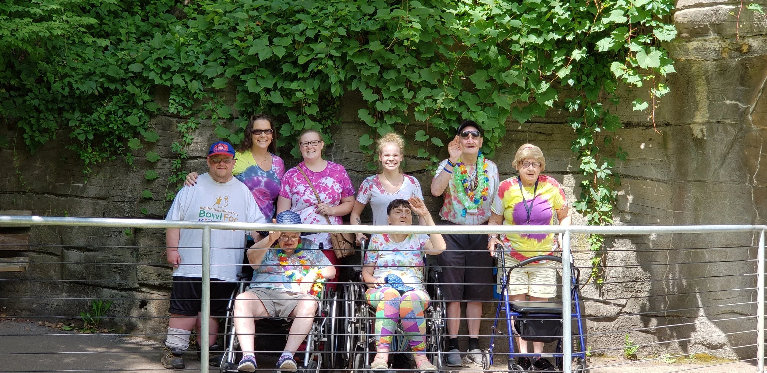 A group of nine people, including elderly women in wheelchairs, standing in front of a stone wall covered with green ivy, posing for a photo on a sunny day.