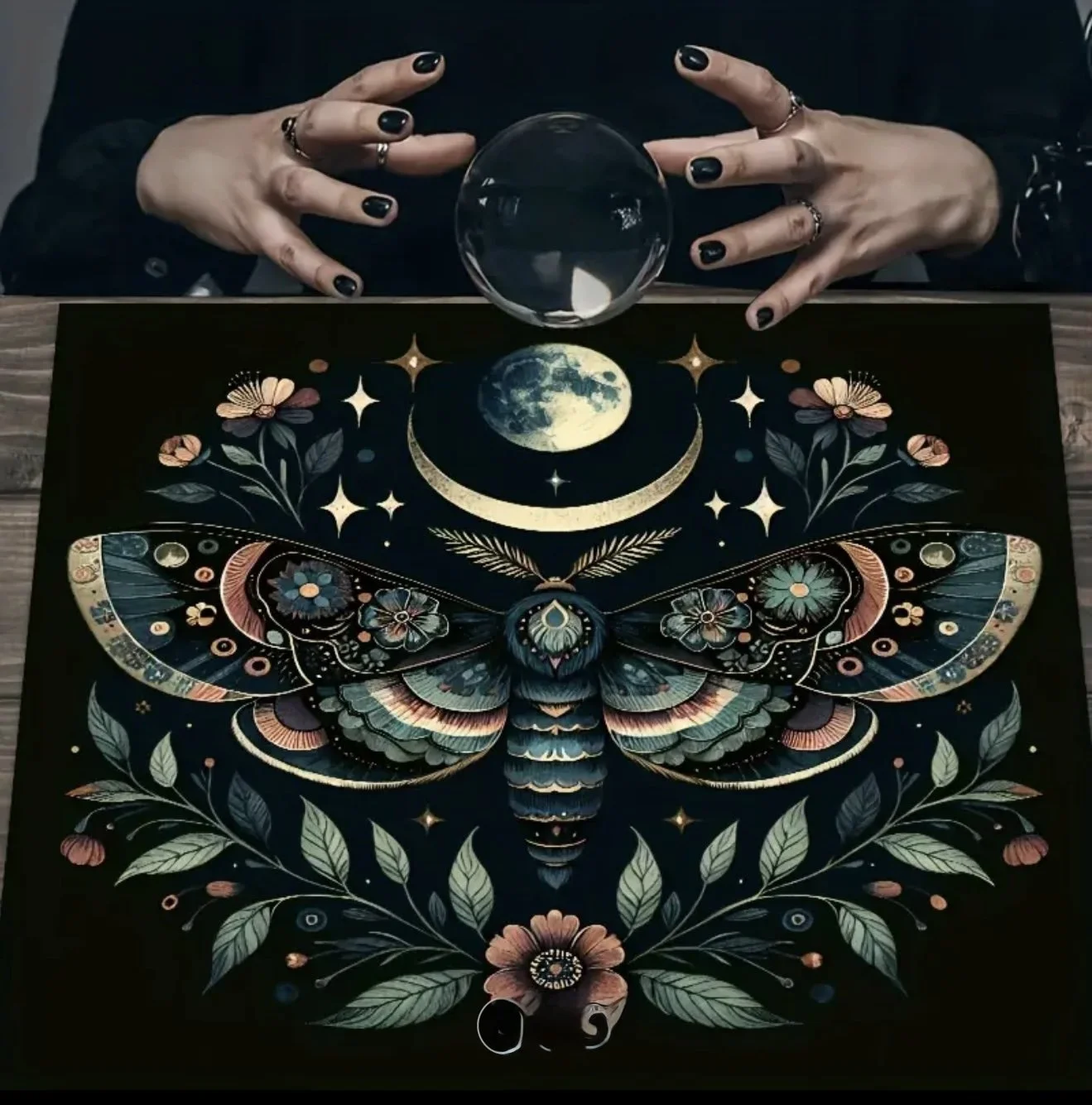 A Tarot card with detailed artwork of a large moth, moon, stars, flowers, and leaves, on a dark background. A crystal ball is placed above the card, with two hands with black painted nails and rings resting on either side.