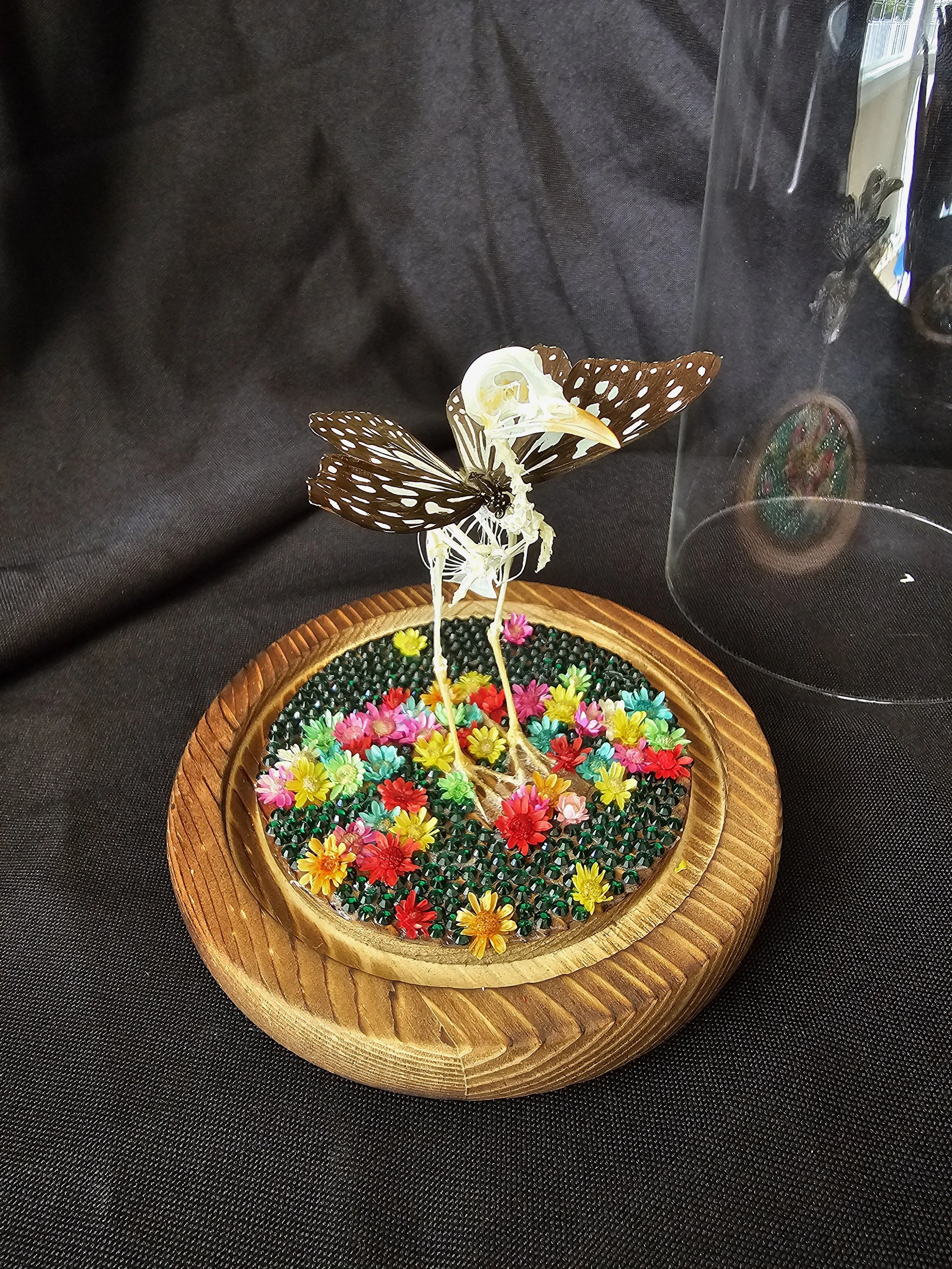 Decorative display with a skeletal bird with butterfly wings and a plastic butterfly perched on a bed of colorful artificial flowers in a wooden circular container.