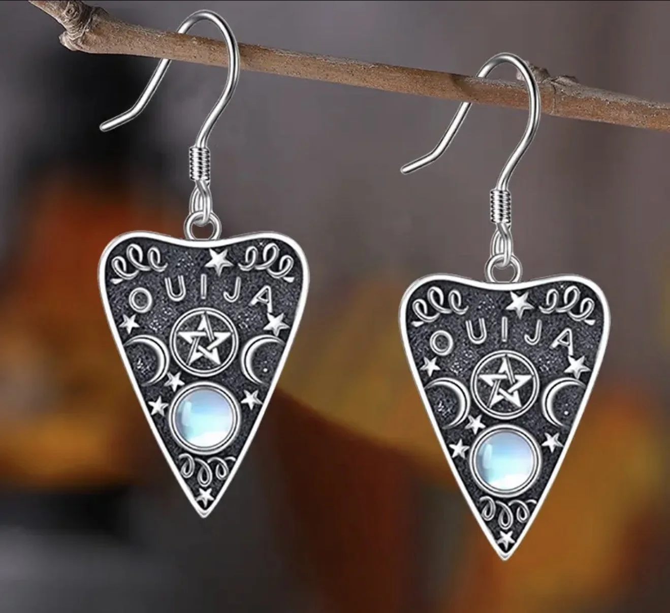 A pair of jewelry earrings shaped like guitar picks with the word 'Ouija', stars, crescent moons, and a central gem on each, hanging from a branch.