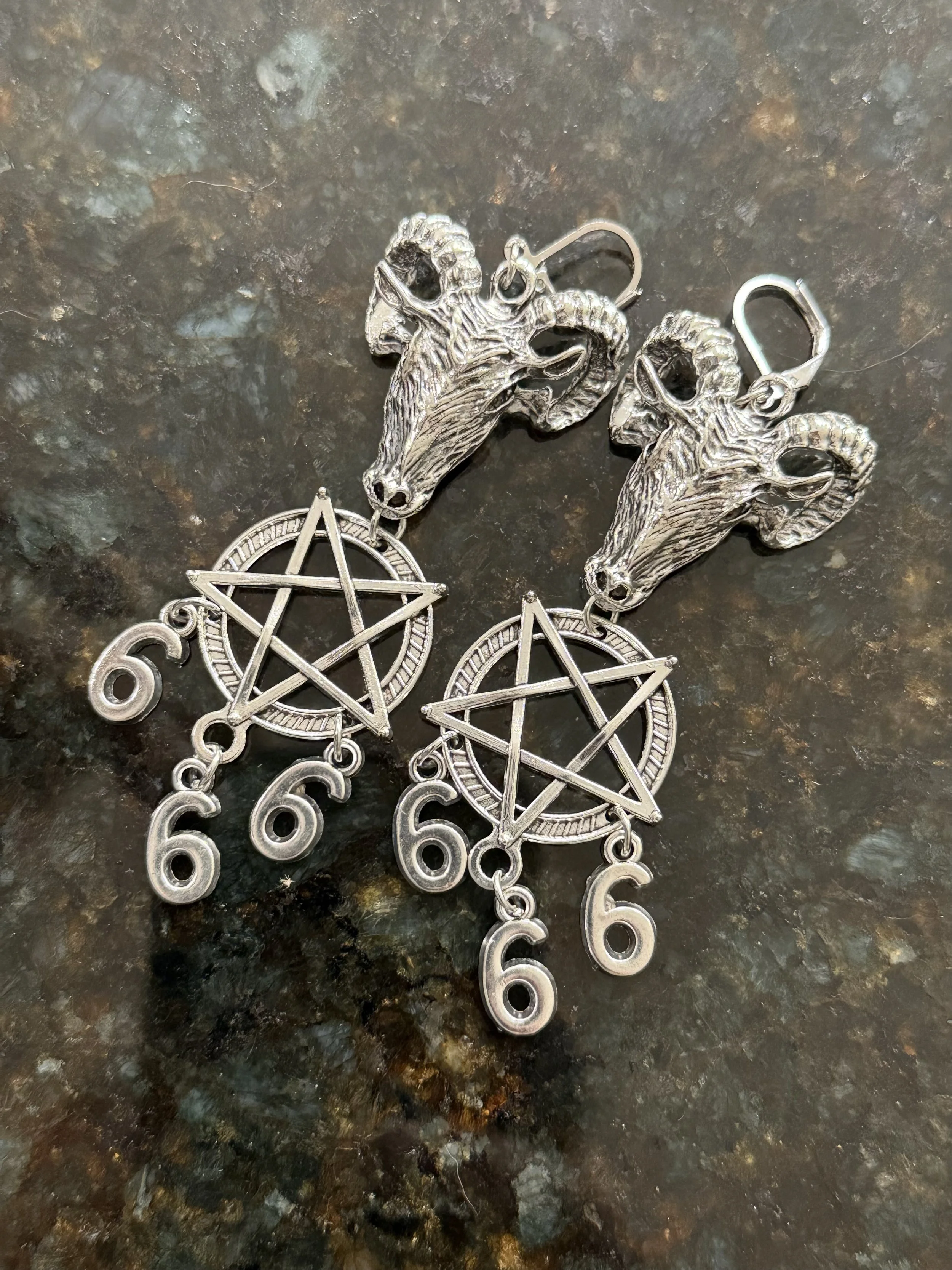 A pair of silver earrings featuring ram's head designs, pentagrams, and the numbers six hanging below.