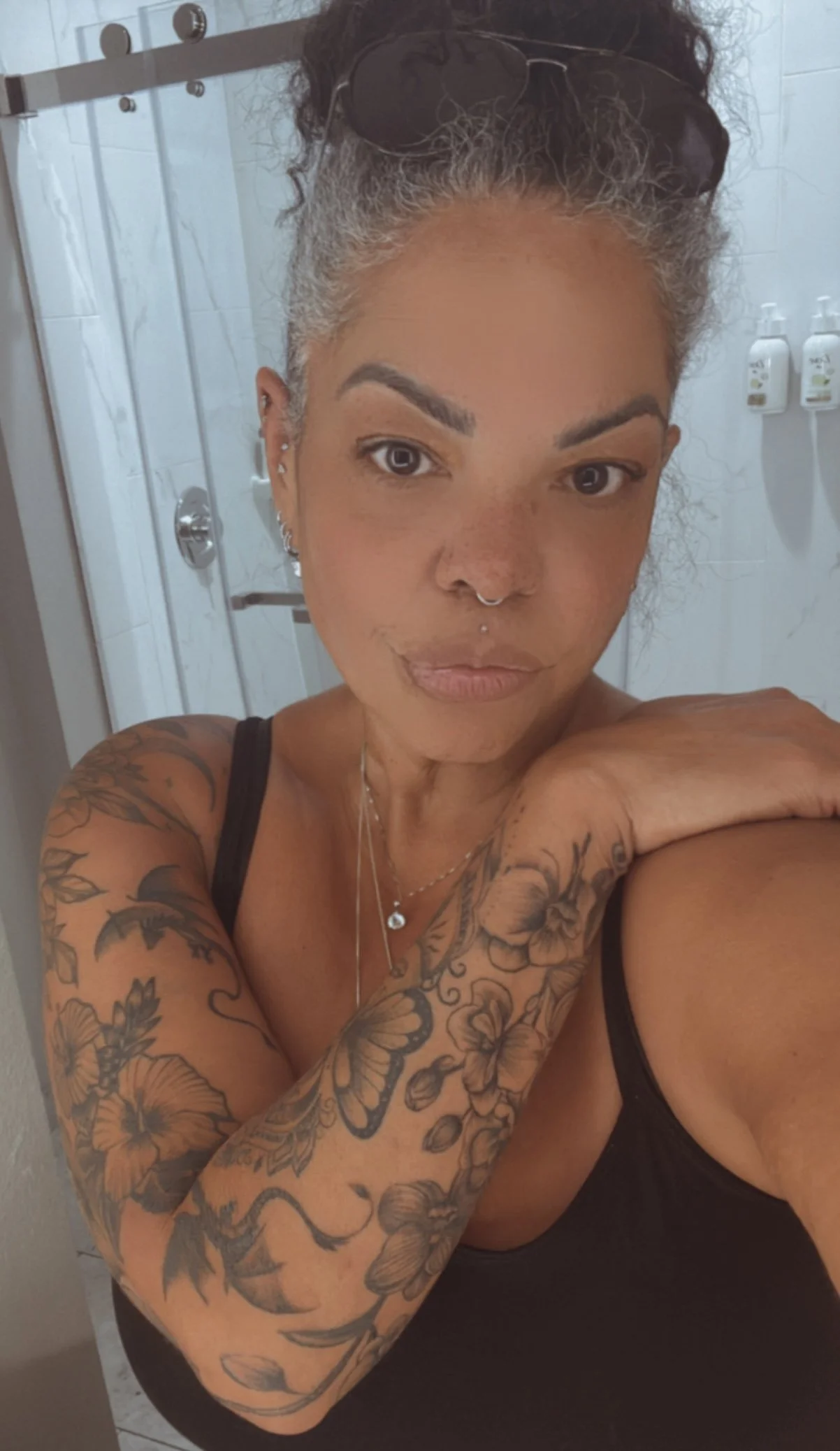 A woman taking a selfie in a bathroom mirror, with tattoos on her left arm, wearing a black tank top, and accessorized with a necklace, earrings, nose ring, and sunglasses on her head.