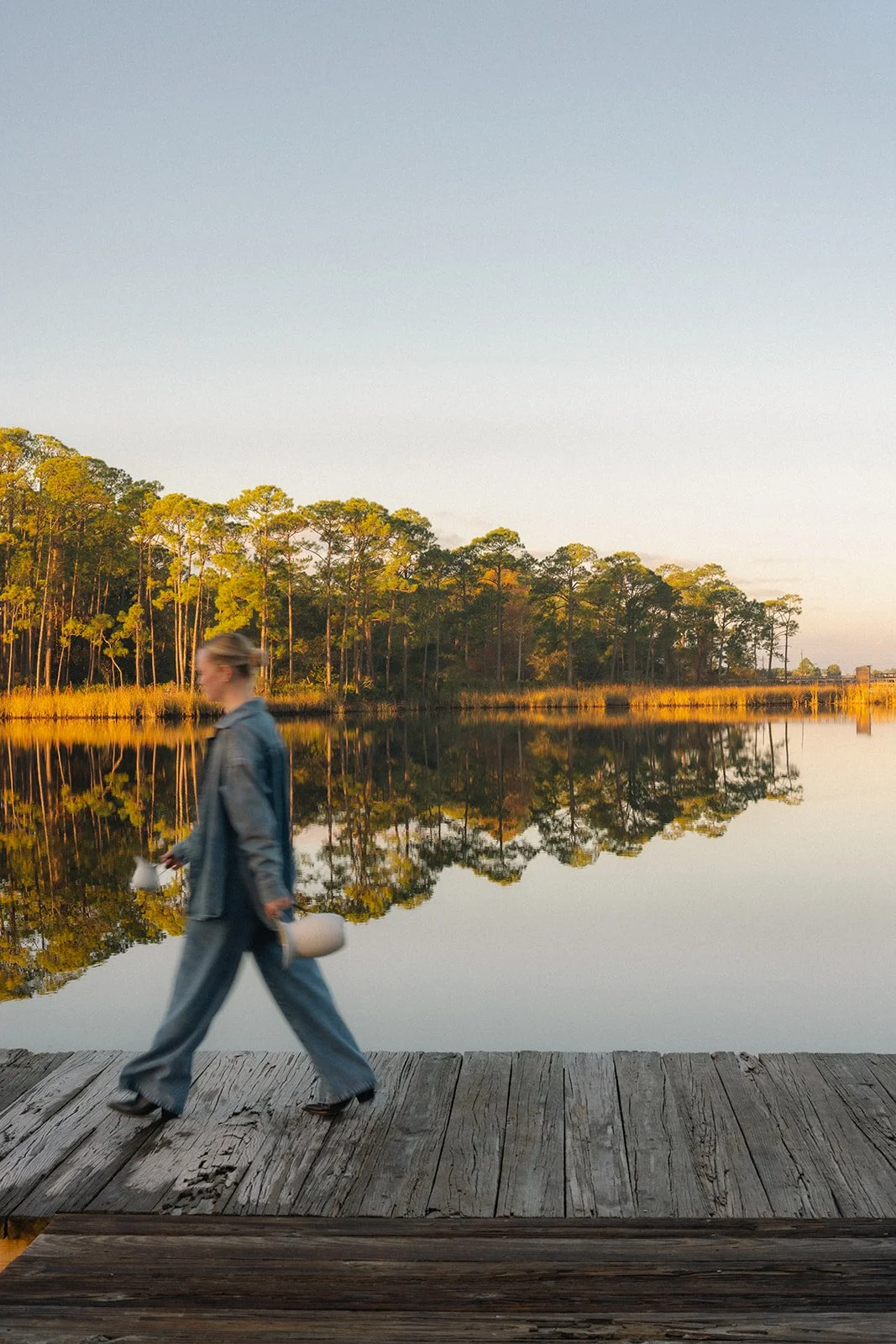A blurred woman walking on a wooden dock along a reflective lake with trees and a clear sky in the background at sunrise.