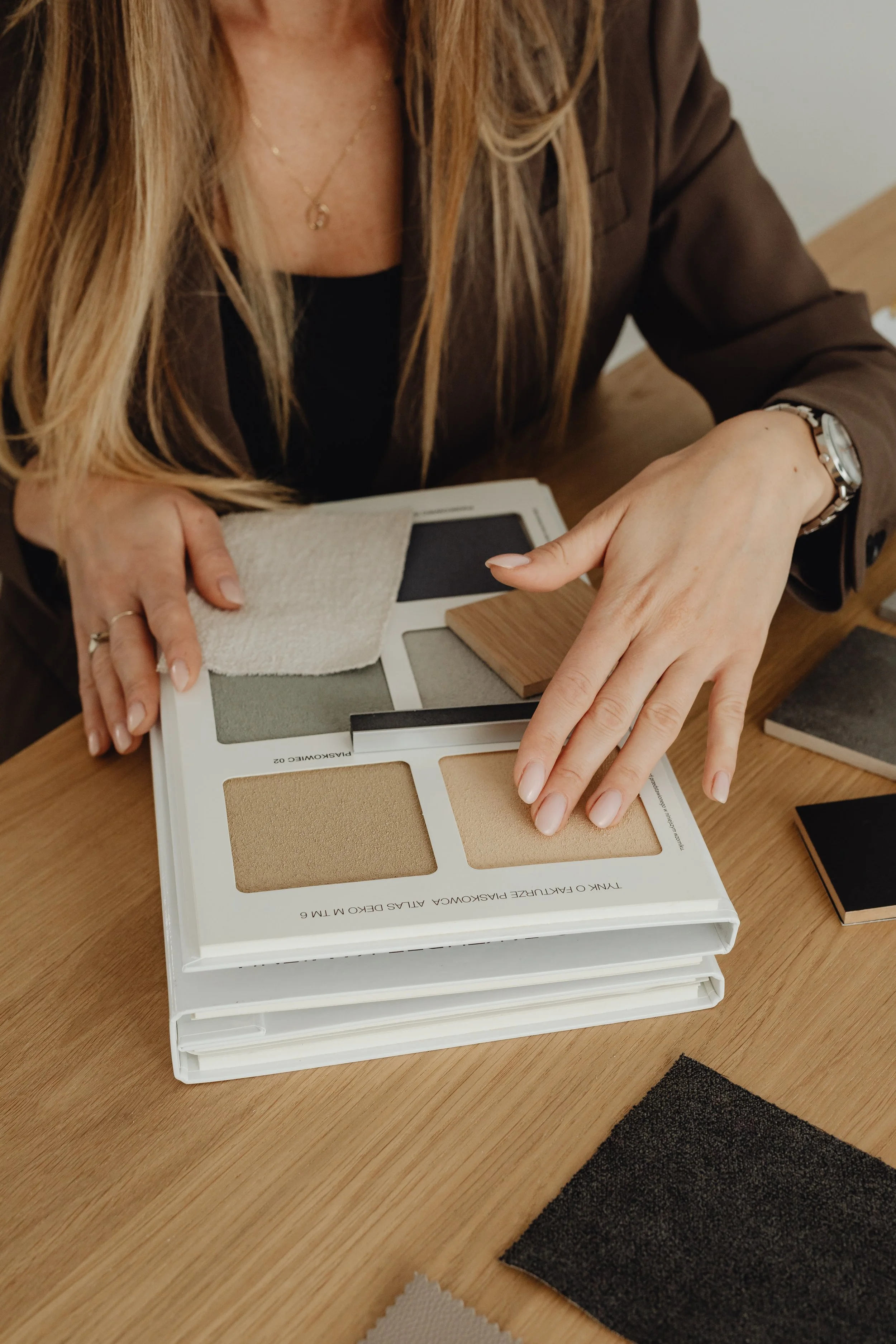 A woman is sitting at a wooden table, holding a color palette book with various fabric and material samples, likely discussing interior design or home decor.