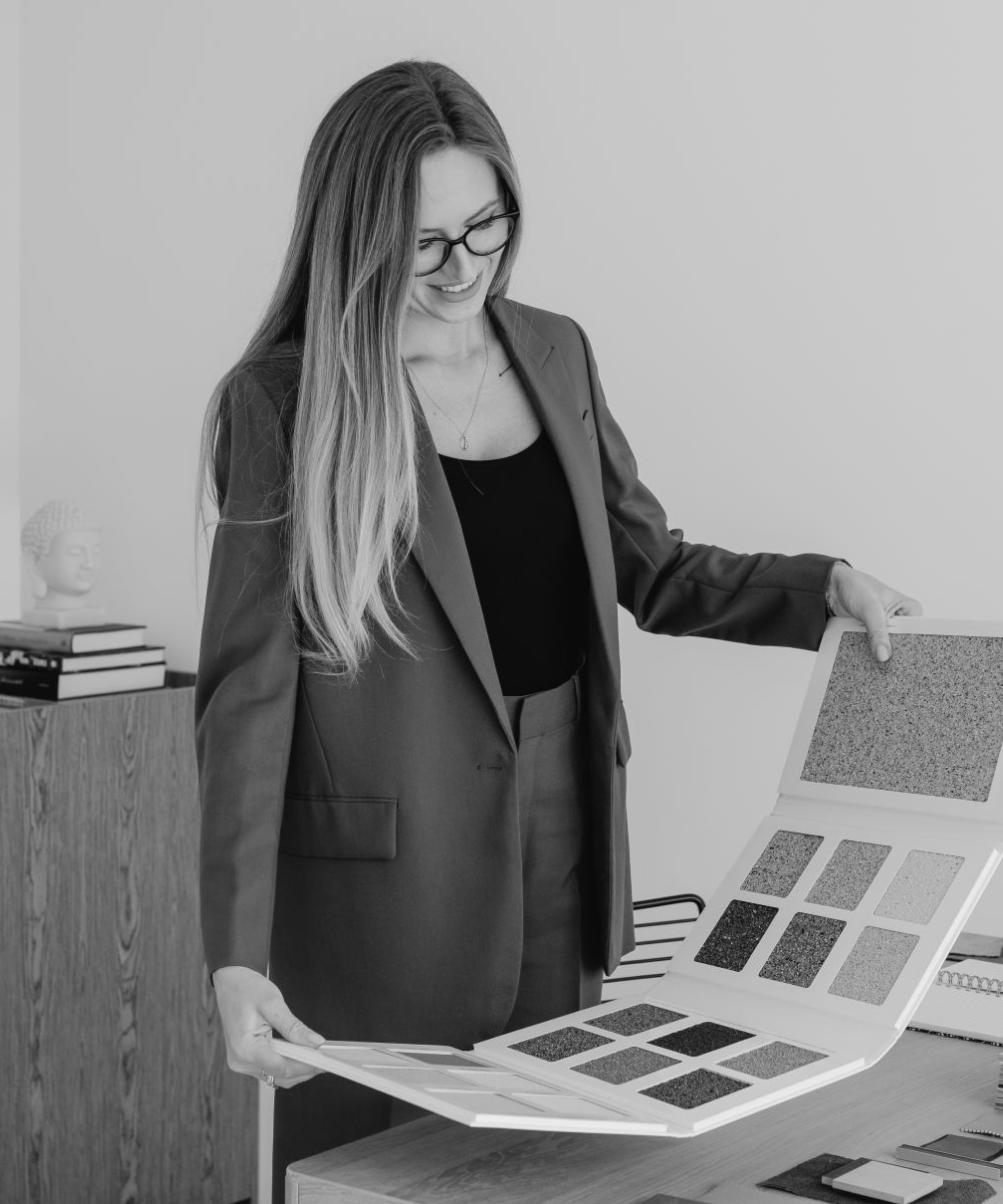 A woman with long hair and glasses in a business suit looking at a color sample book in an office setting.