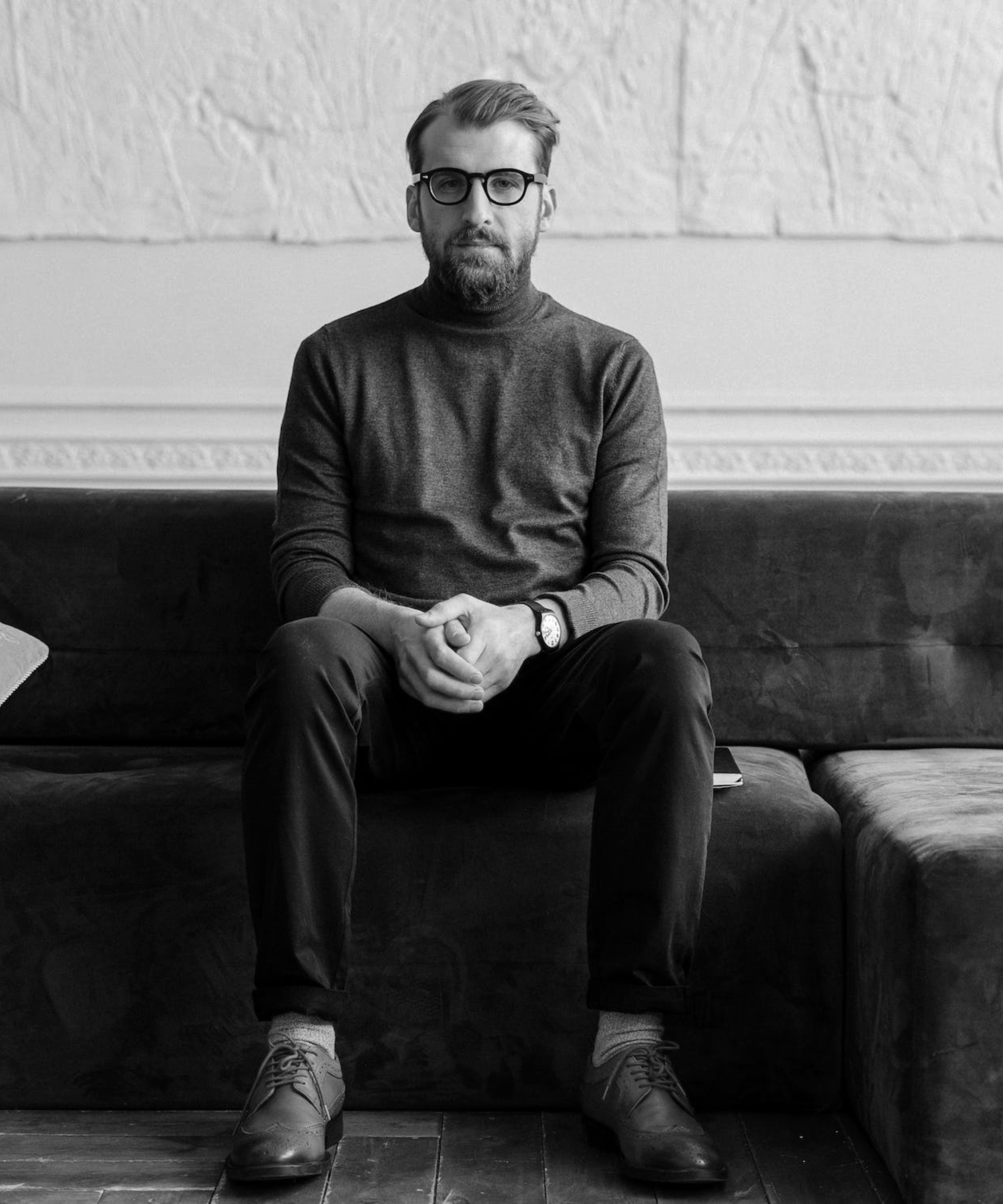 A man with glasses and a beard sitting on a dark sofa in a black-and-white photo, wearing a turtleneck, pants, and shoes.