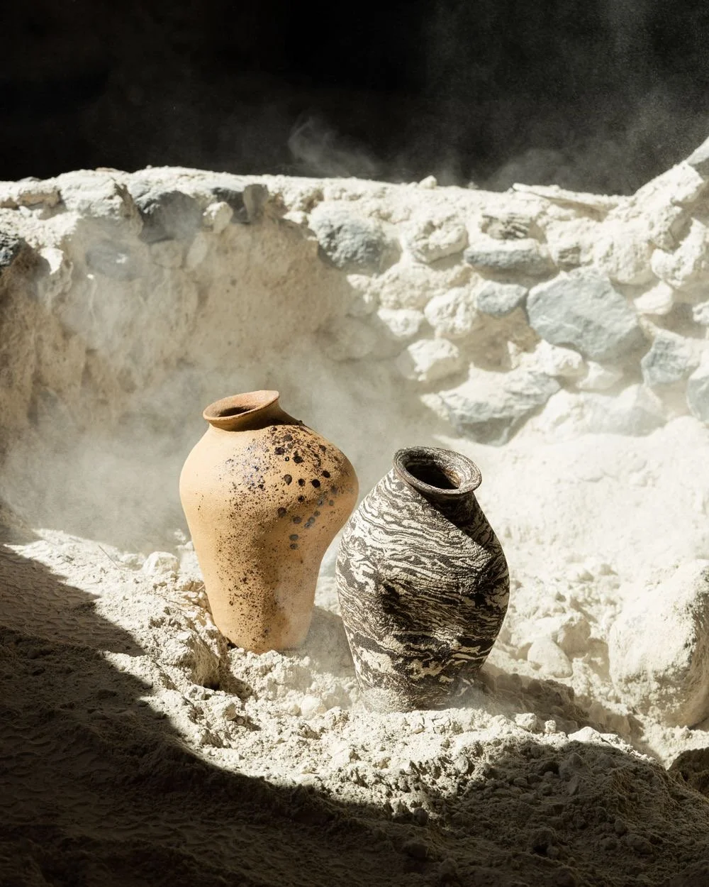 Two ancient pottery vases, one beige with black speckles and one black and white marble patterned, are placed on white sandy ground with a white stone wall behind them, emitting steam or smoke.