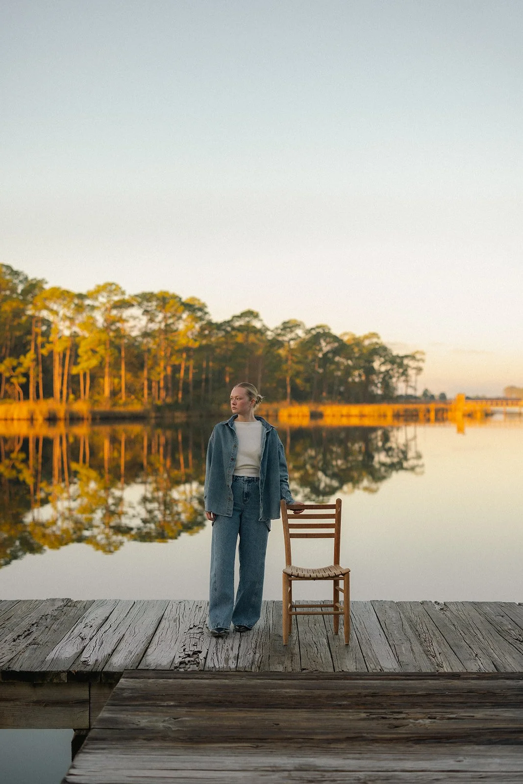 A woman standing on a wooden dock by a calm lake with trees in the background during sunset, with an empty wooden chair beside her.