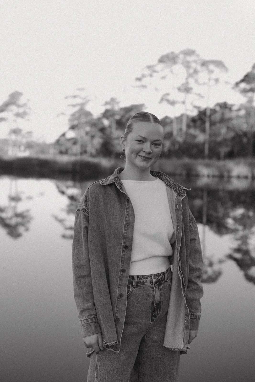 A young woman standing outdoors near a body of water, smiling at the camera, with trees in the background, in black and white.