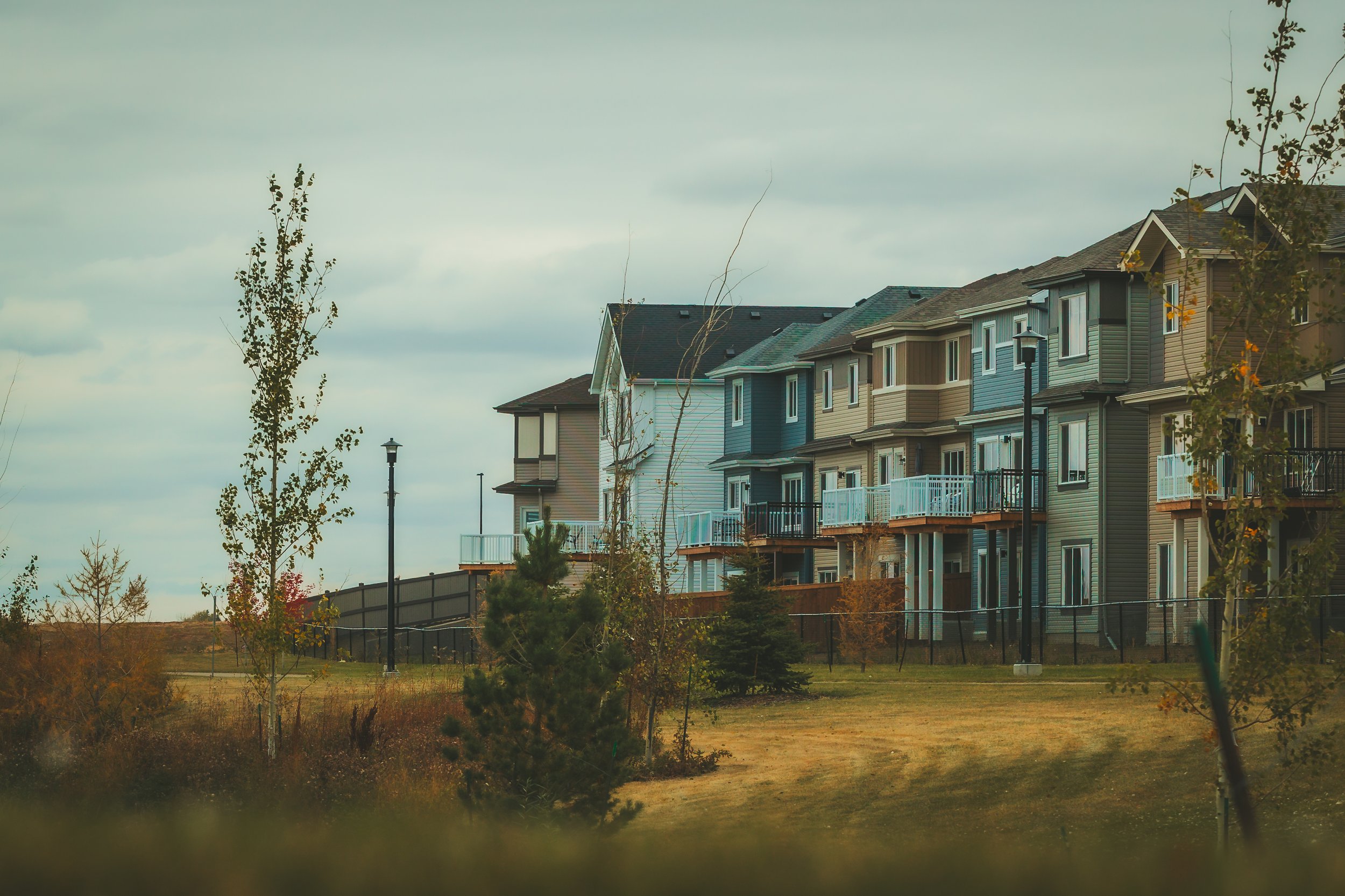 Multi-story residential apartment buildings with balconies set above a landscaped area with trees and grass, under a cloudy sky.