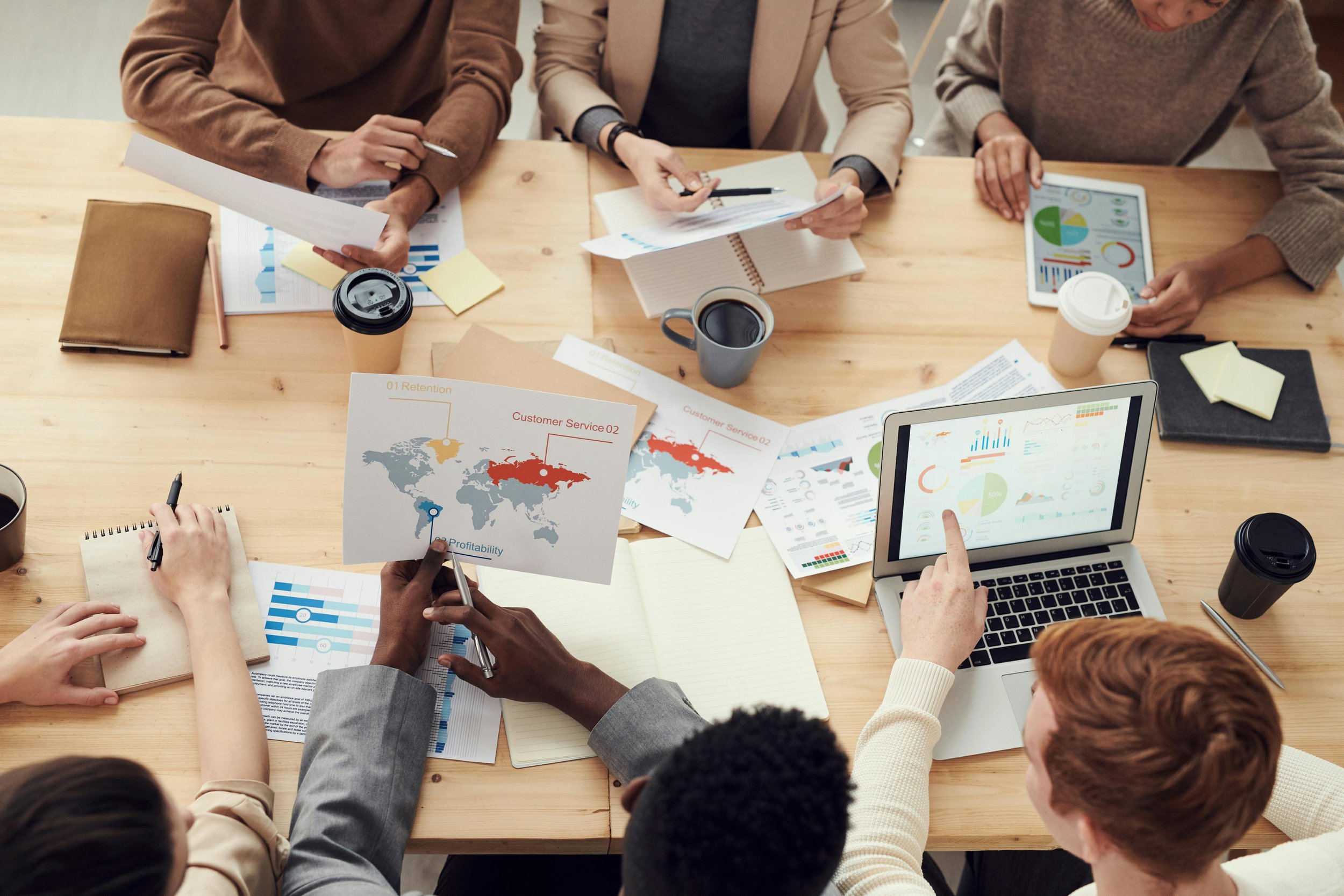 A group of people sitting around a wooden table in a business meeting, reviewing charts, graphs, and a laptop displaying data visualizations.