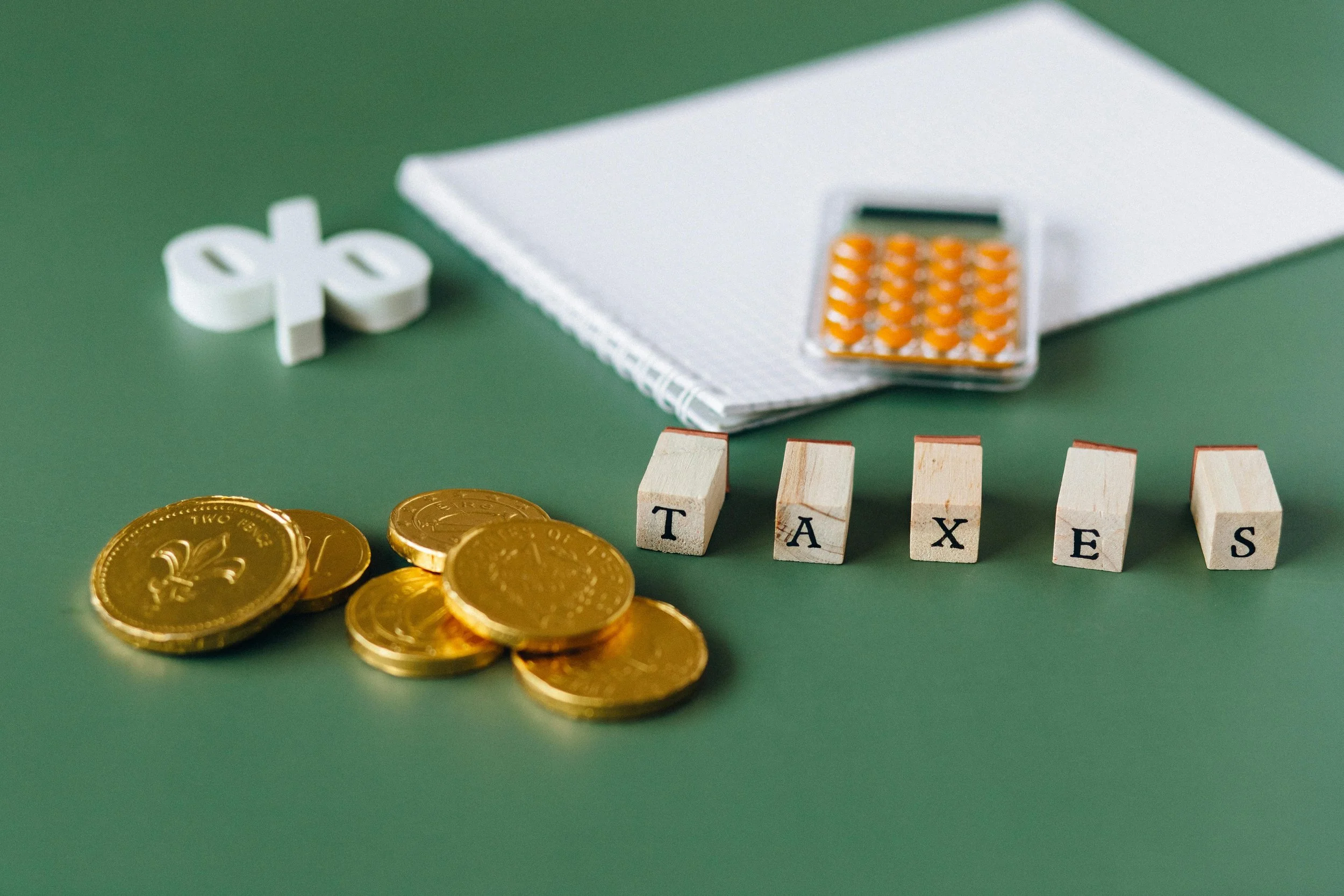 Coins, dice, a notepad, a calculator, and wooden blocks spelling 'TAXES' on a green surface.