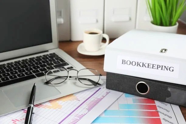 Desk with a laptop, pair of glasses, a pen, a coffee cup on a saucer, a white book labeled 'Bookkeeping' on a black file folder, some colorful charts, and a green plant in the background.
