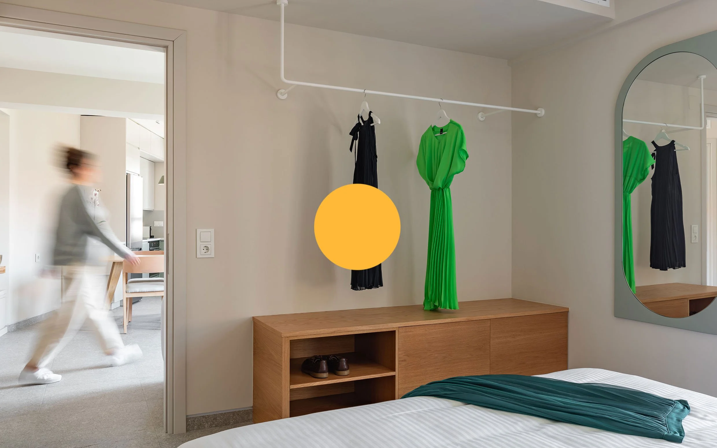 Bedroom with a wooden shelf, a green dress and a black dress hanging on a white clothing rod, a mirror reflecting the clothes, and a woman walking out of the room, all in a calm, modern style.