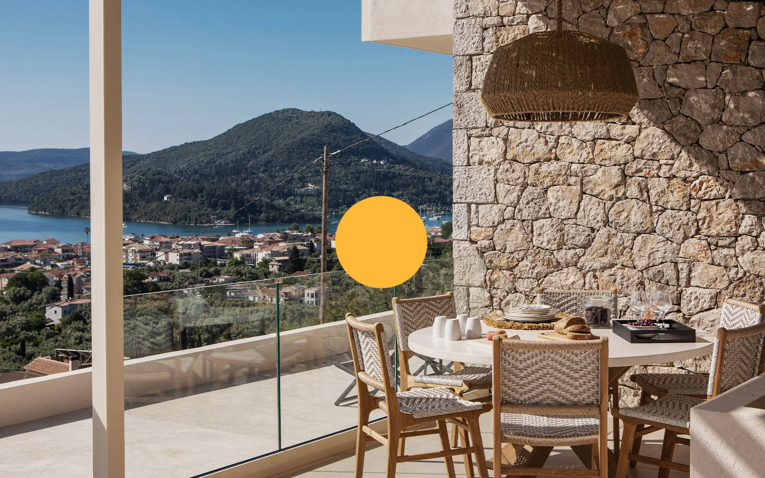 Outdoor dining area with a round table and six chairs, overlooking a scenic view of hills, a lake, and a town, with a stone wall and a woven hanging light