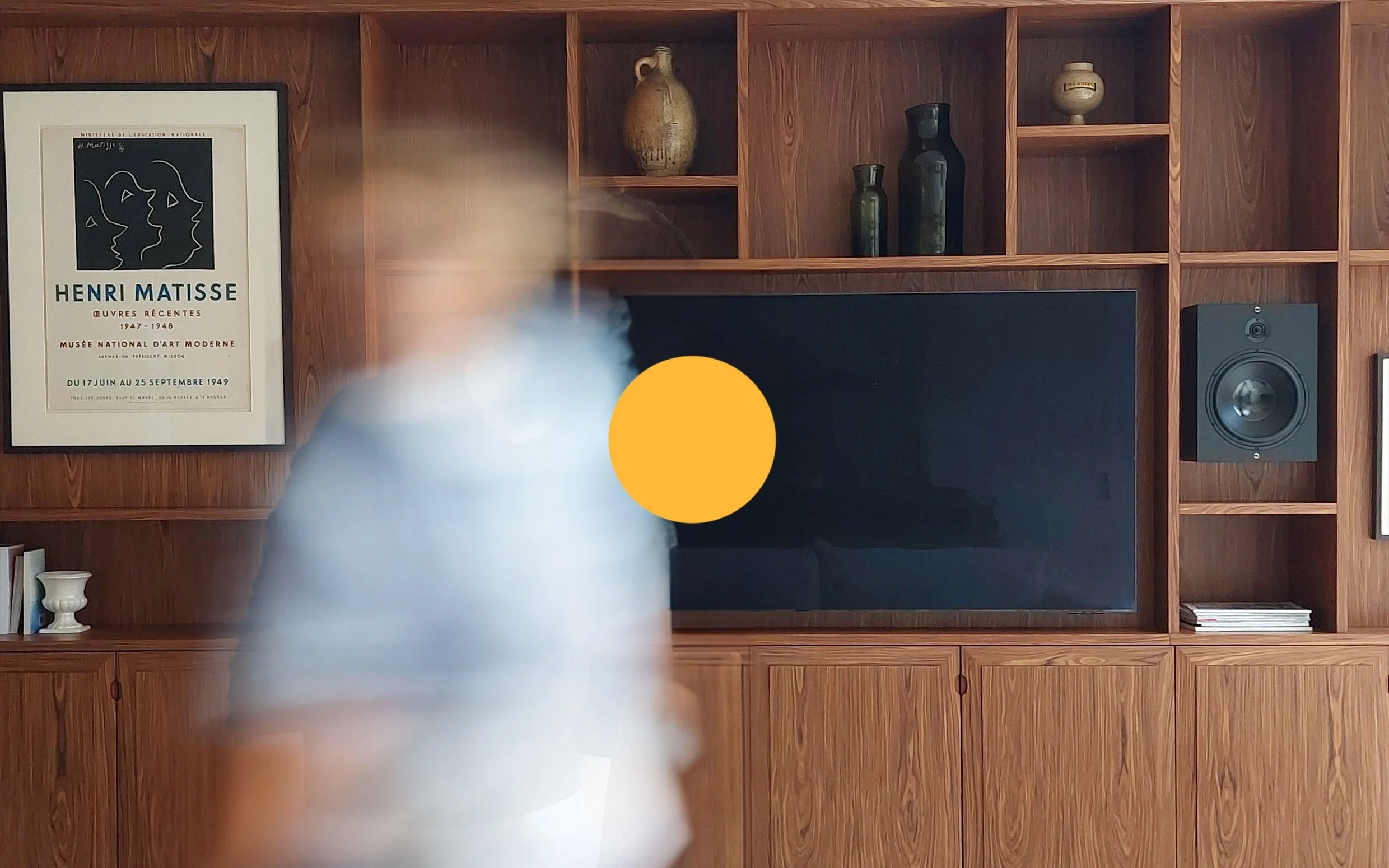 Living room with a large wooden entertainment center, a TV, a speaker, artwork, and decorative vases, with a person blurred in the foreground.