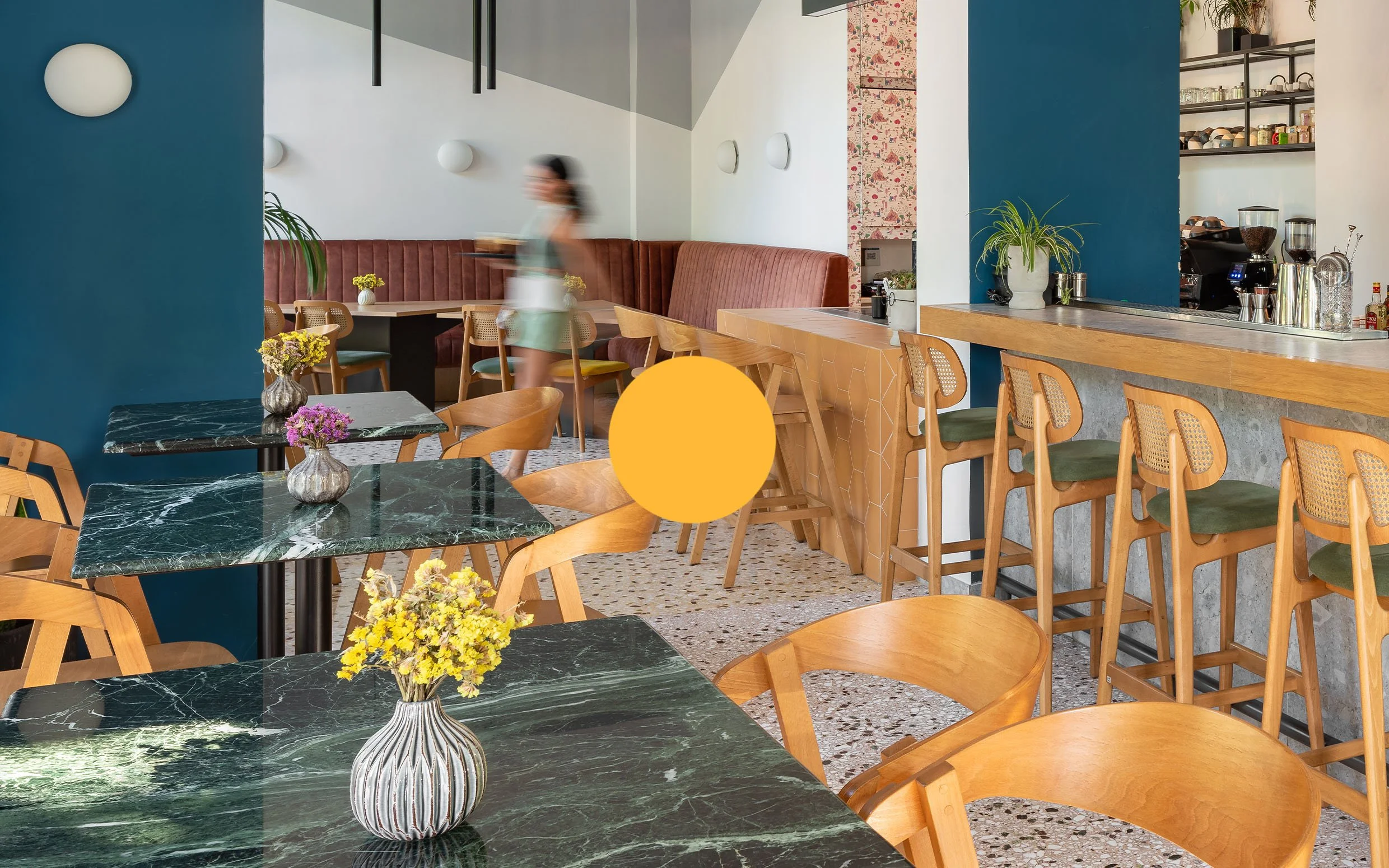 Contemporary cafe interior with green marble tables, wooden chairs, a bar counter, and a booth with red upholstery. Decor includes colorful vases with flowers, potted plants, and modern lighting fixtures.