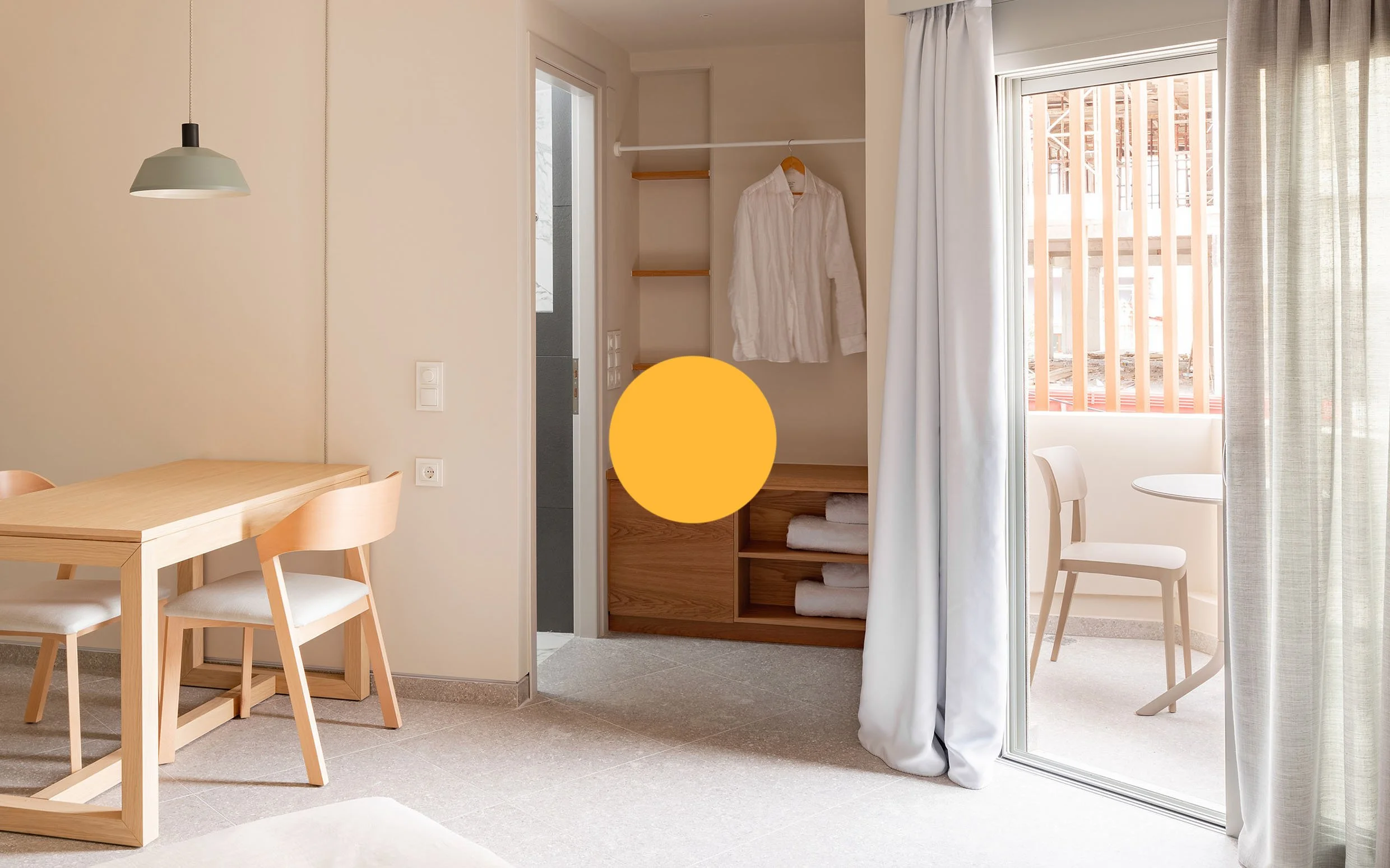 Interior of a modern minimalistic hotel room with a dining table, chairs, open closet with towels and a white shirt, sliding glass door with curtains leading to a small balcony with a chair.