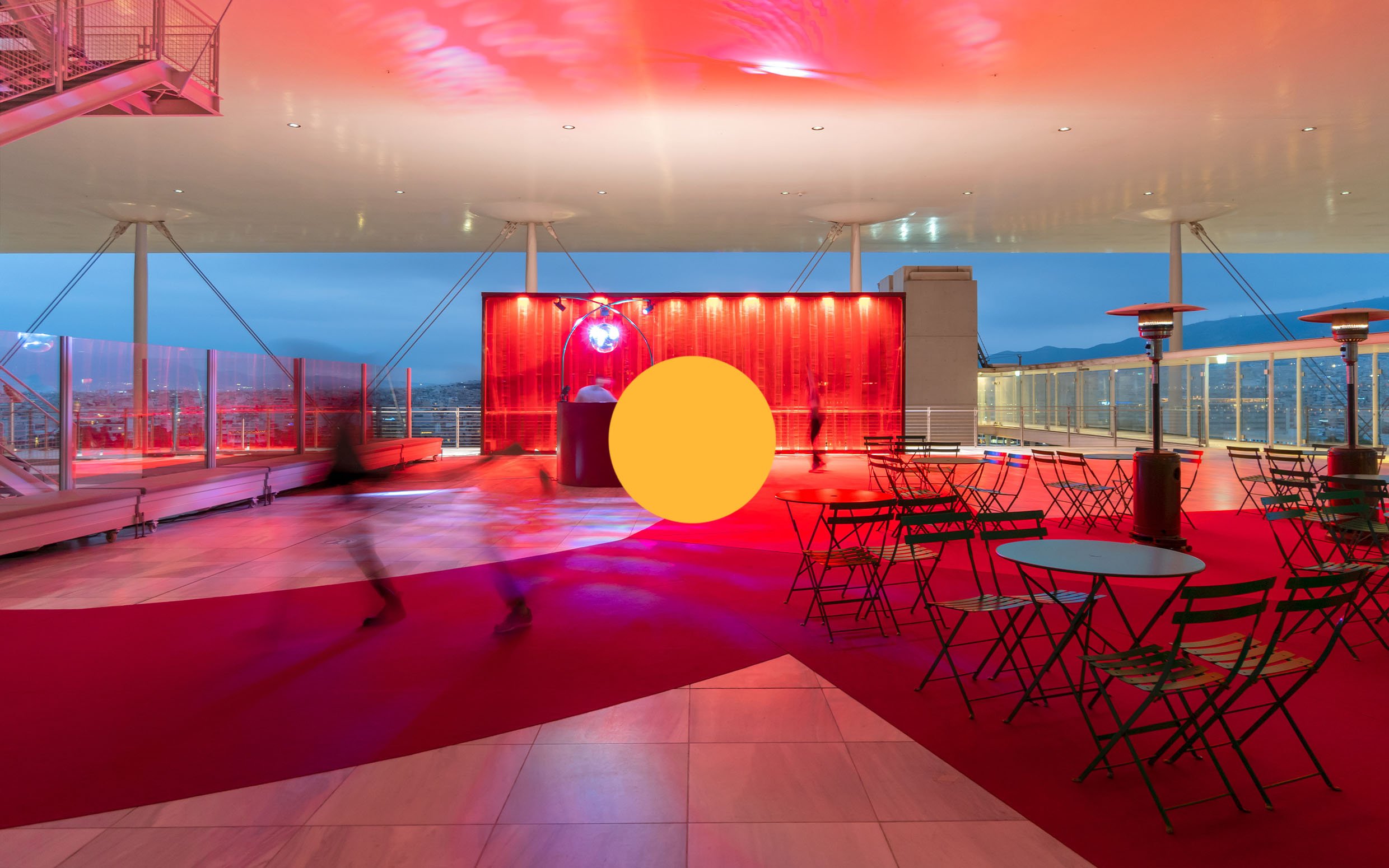 Rooftop lounge area with red lighting, empty tables and chairs, and blurred figures walking by, overlooking cityscape at dusk.