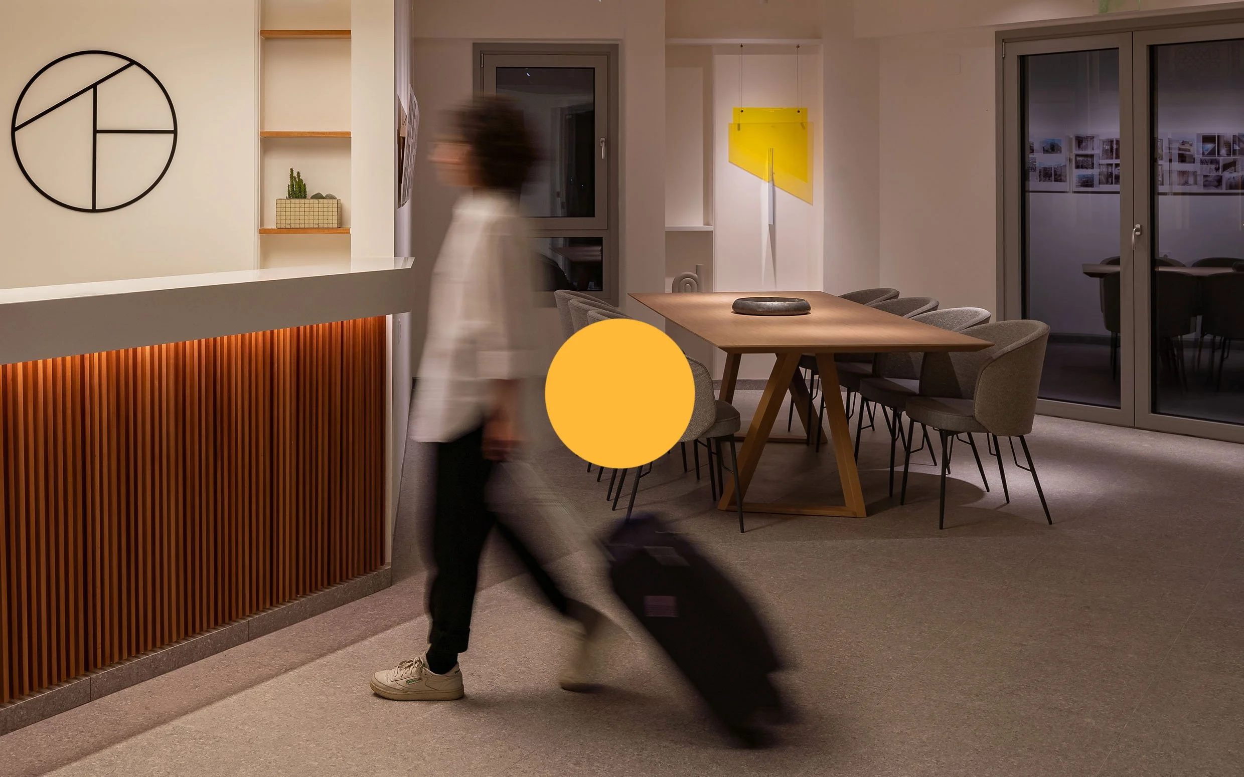 Person walking through a modern, well-lit lobby area with a wooden reception desk, a large dining table with chairs, and large windows at night.