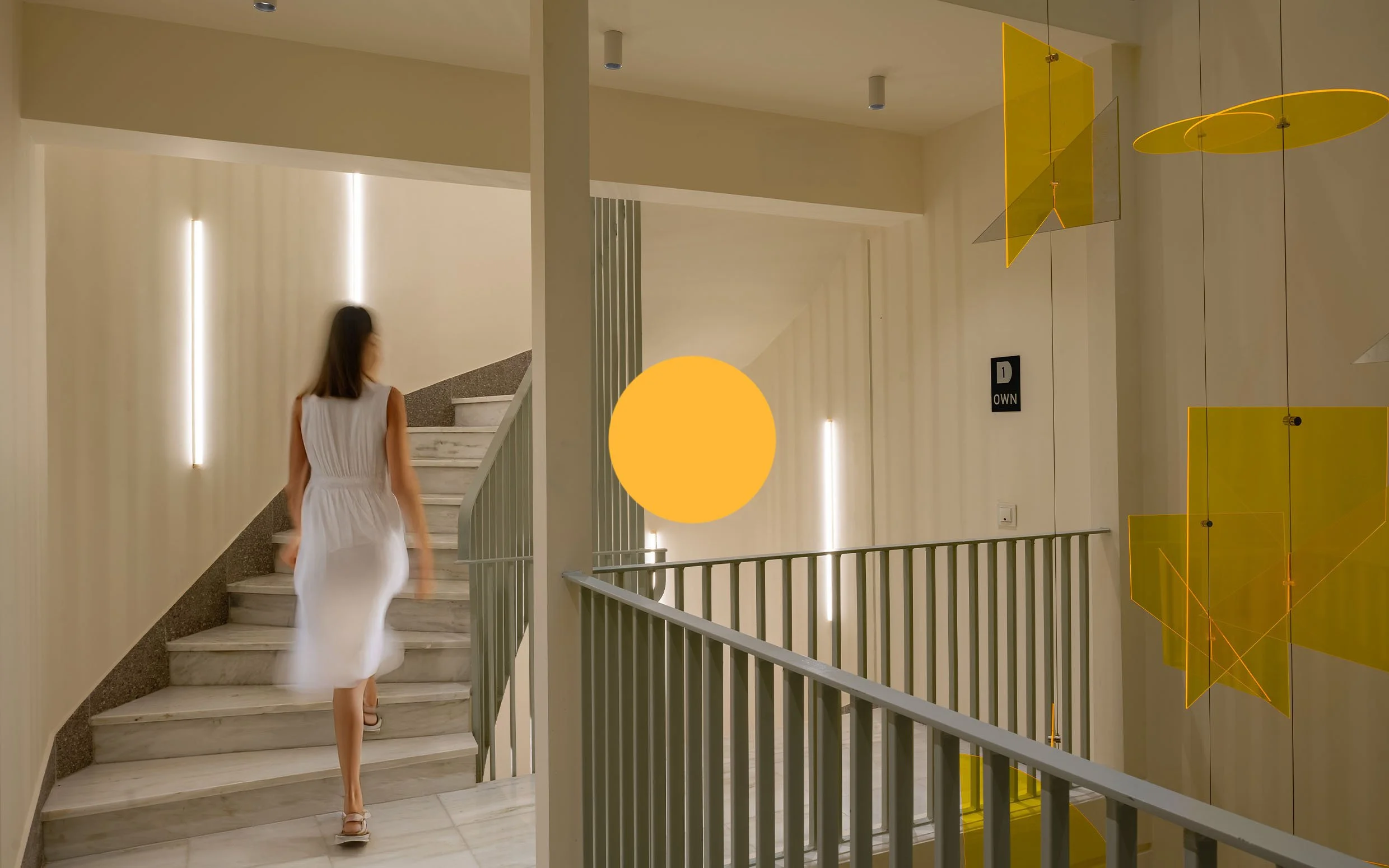 Woman in white dress walking up marble stairs in modern, well-lit interior with vertical light fixtures and yellow decorative hanging panels.
