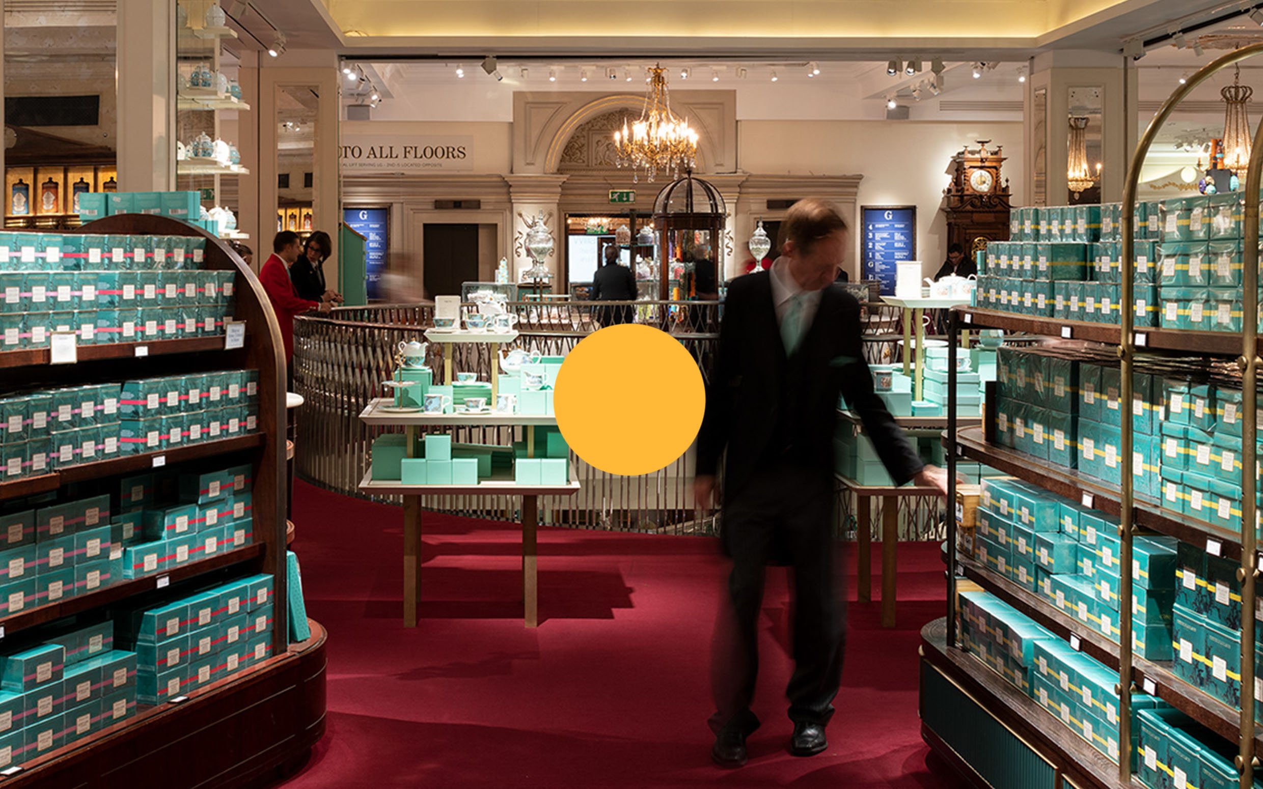 A person in a black suit shopping in a store with shelves of turquoise boxes inside a grand, elegant interior with chandeliers and antique decor.