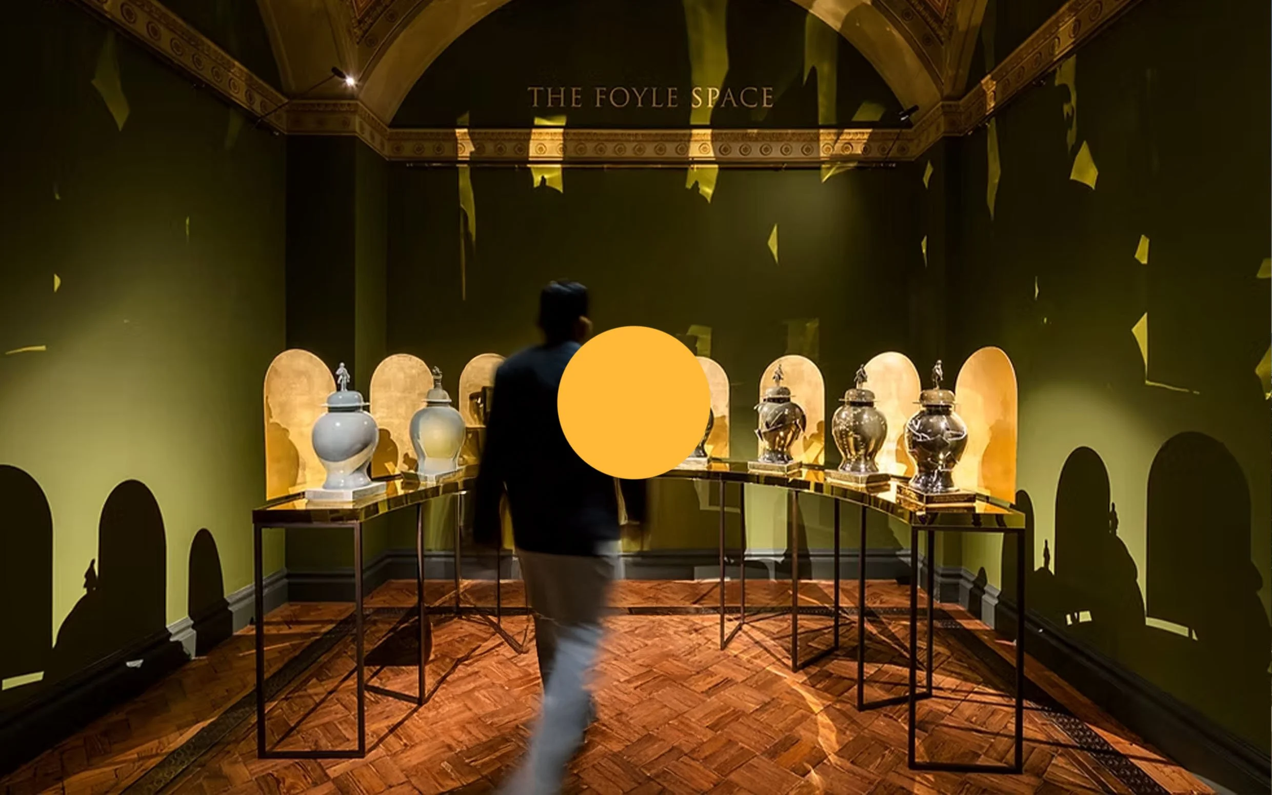 An art gallery exhibit titled 'The Foyle Space' features a man walking past a circular display table with large, ornate jars or urns. The exhibit is in a dimly lit room with green walls, arched niches, and golden accents, casting shadows on the walls