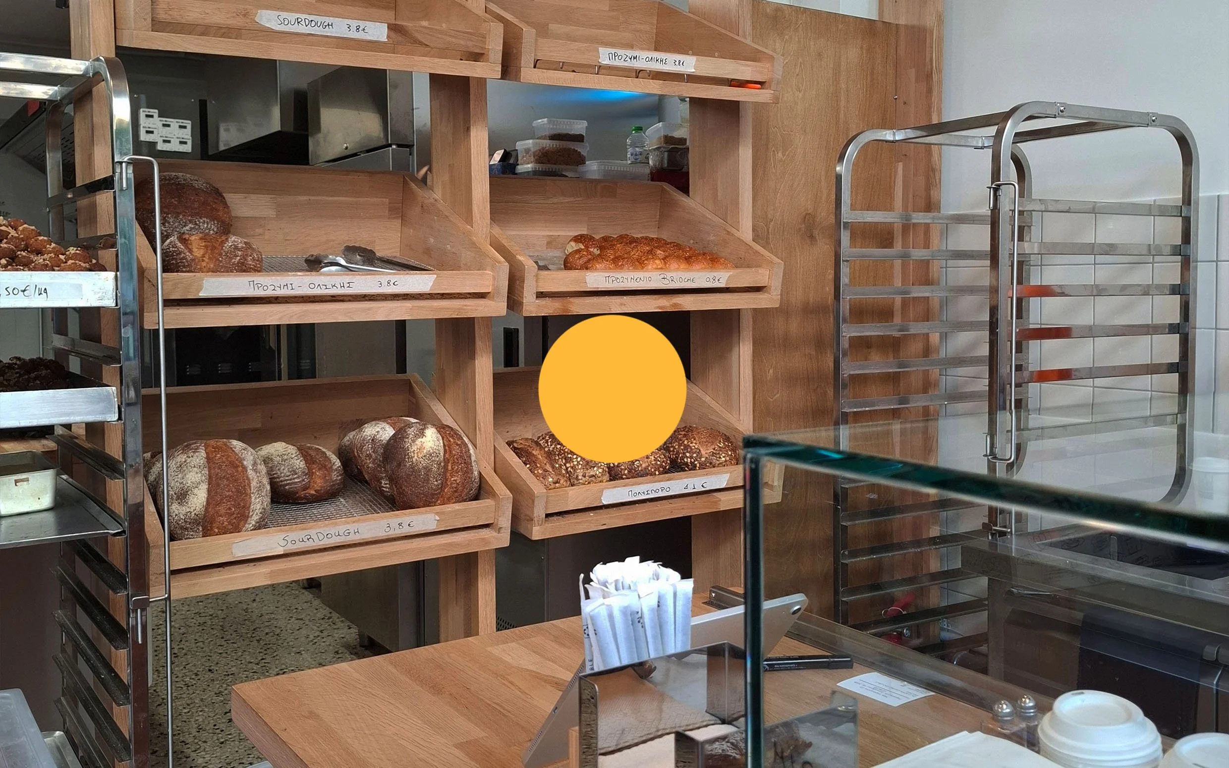 Wooden shelves holding various types of bread, with handwritten labels indicating prices; a metal rack with bread slices and loaves; a glass display case in the foreground; a wooden counter with napkins and utensils.