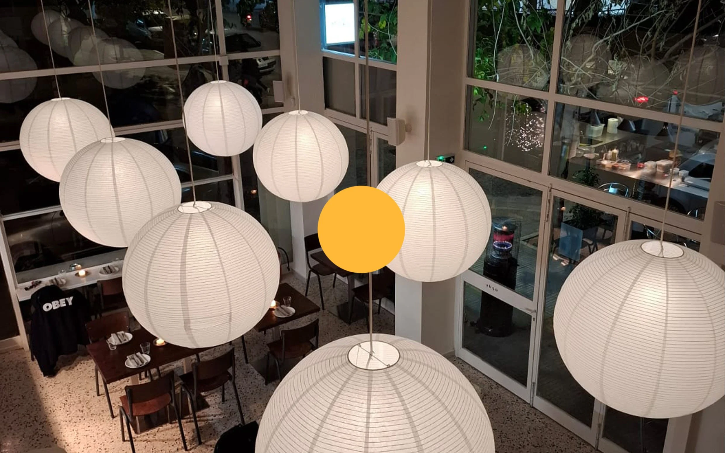 Interior of a restaurant with large white paper lanterns hanging from the ceiling, tables set with napkins and glasses, and a large window showing parked cars and outside street scene at night.