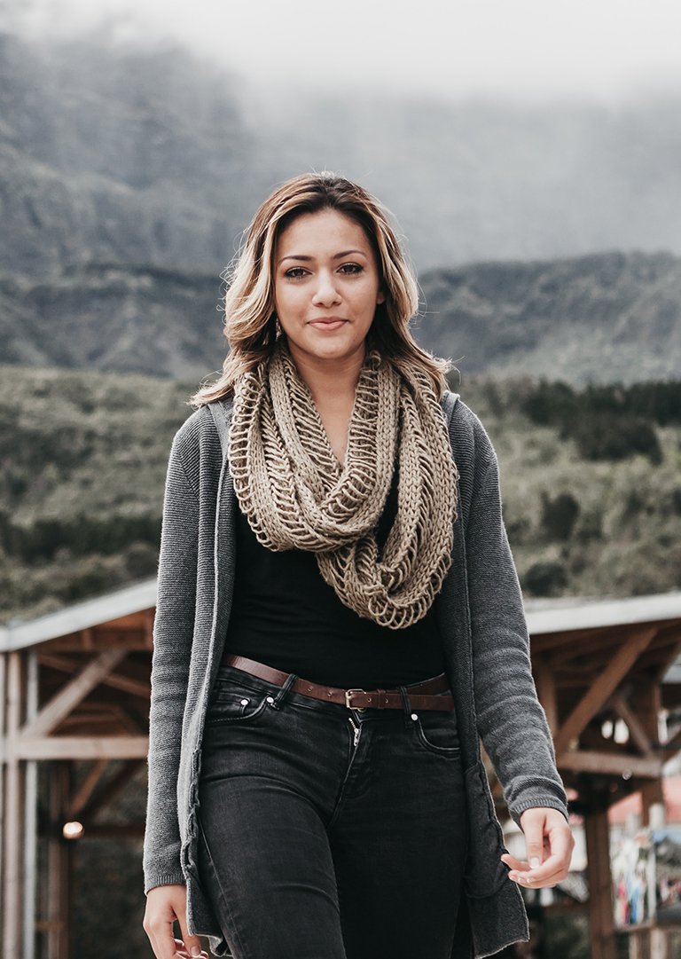 A woman standing outdoors on a cloudy day with mountains in the background, wearing a gray coat, beige scarf, black shirt, and black jeans.