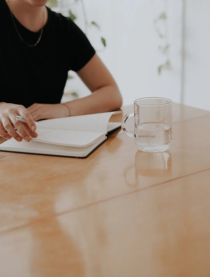 Person with short hair sitting at a wooden table, reading a notebook, with a glass of water nearby.