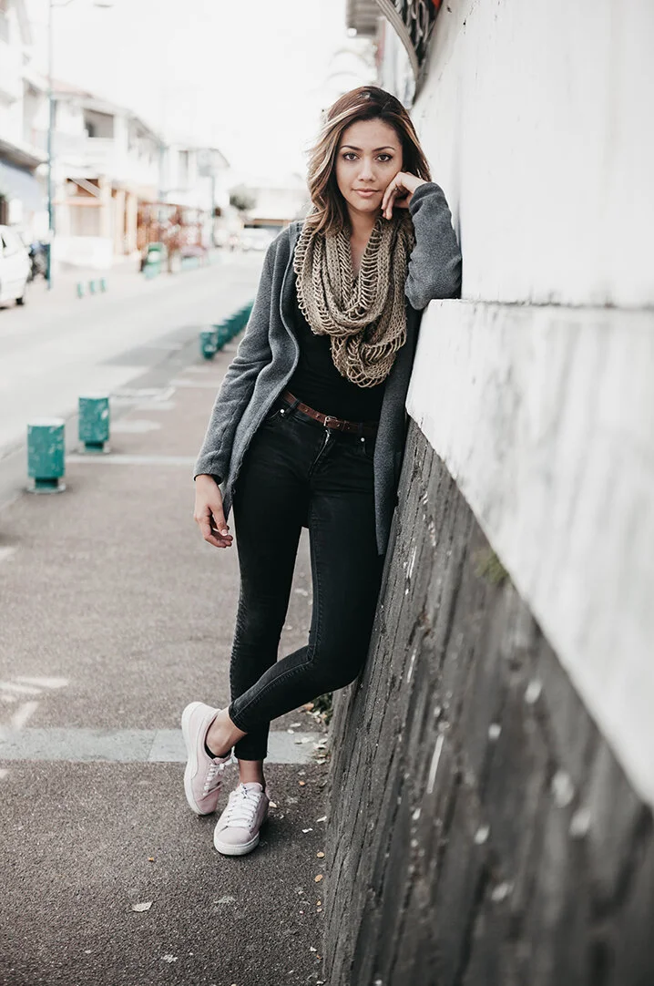 A young woman in casual attire leaning against a wall on a city street, wearing a gray coat, black jeans, a beige scarf, and pink sneakers.