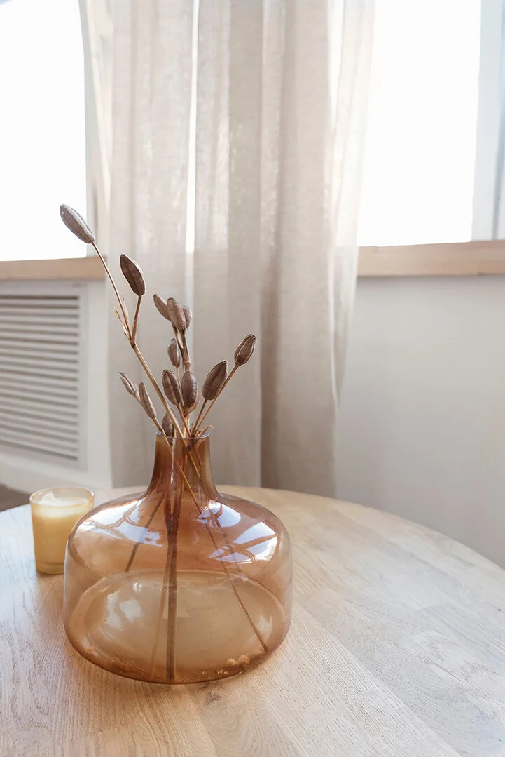 A brown glass vase with dried seed pods and stems on a light wood table, with a small candle and beige curtains in the background.