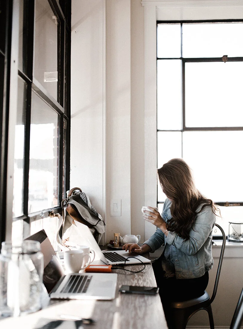 Woman sitting at a wooden table with laptops, coffee cups, and a backpack, working or studying near a large window with sunlight