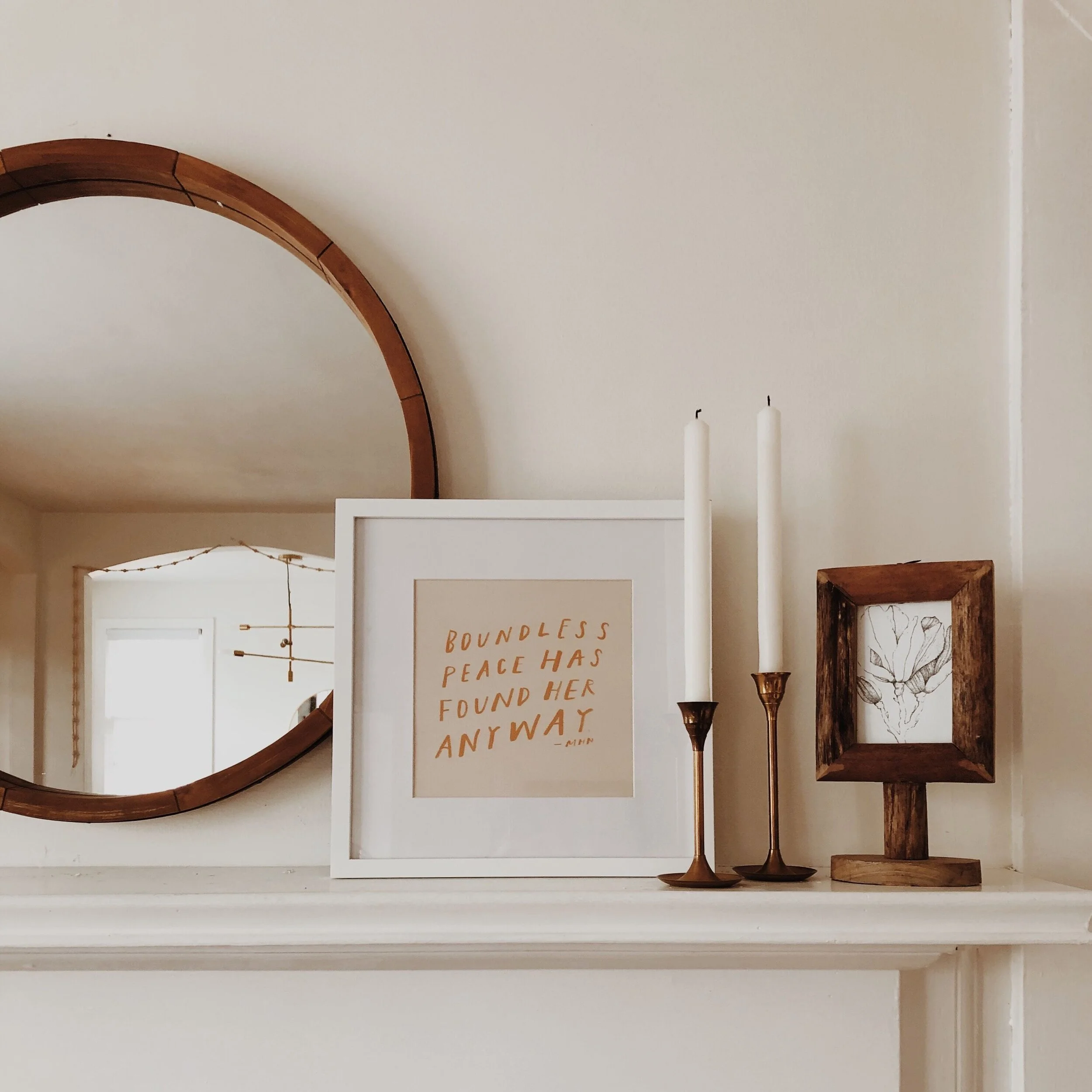 Decorative mantel with a round wooden mirror, framed quote print, two brass candle holders with white candles, and a wooden frame with a botanical drawing inside.