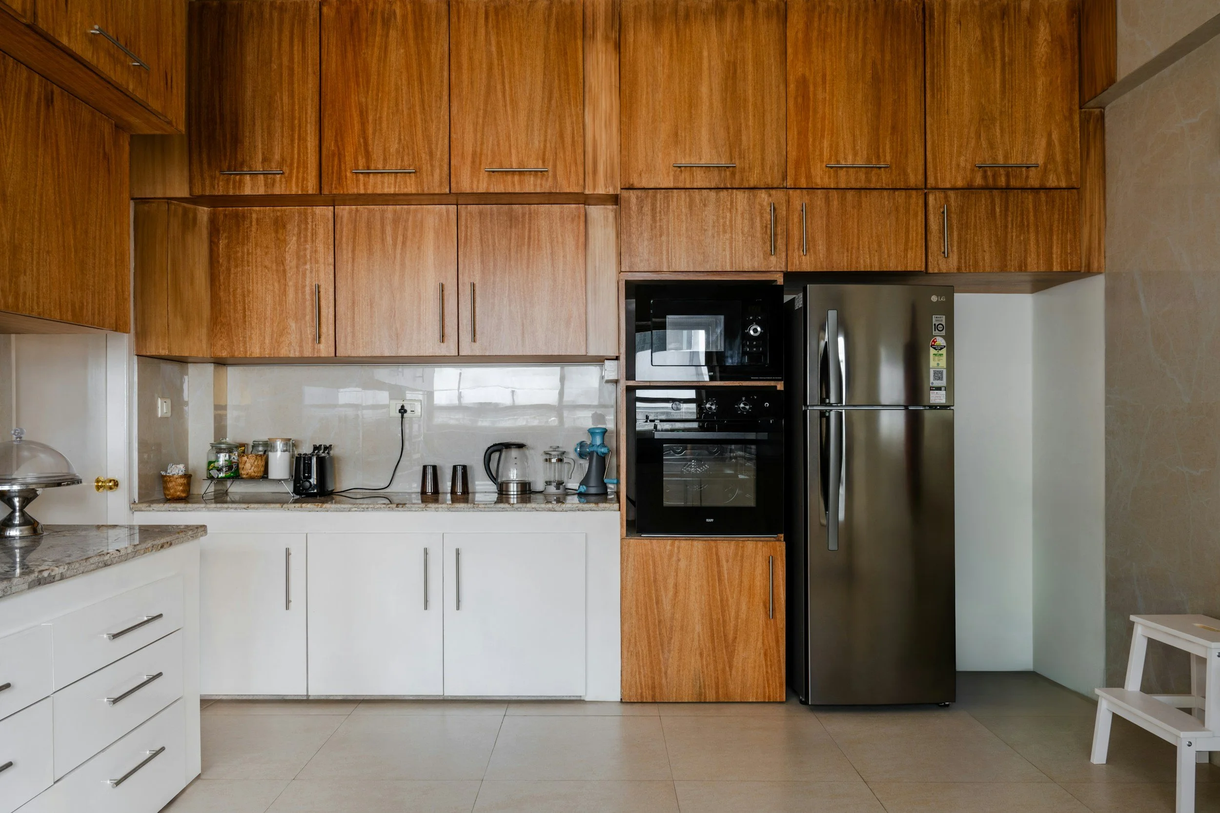 Modern kitchen with wooden upper cabinets, white lower cabinets, stainless steel refrigerator, built-in microwave and oven, and beige tiled floor.