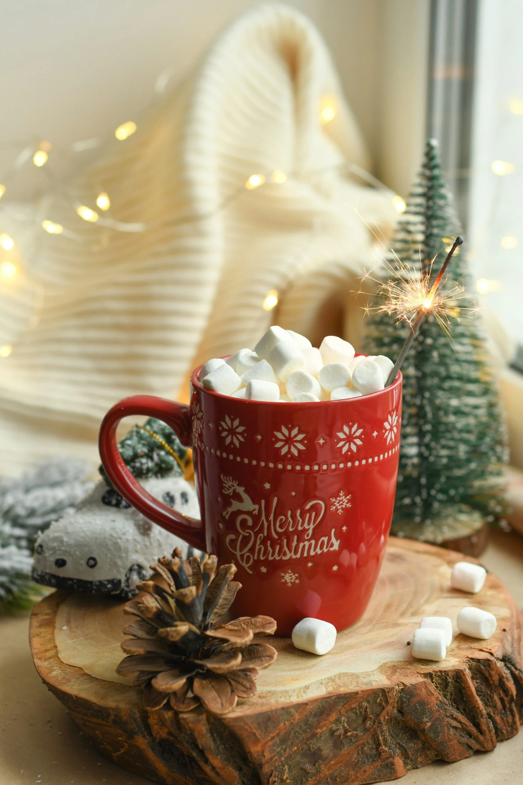 A red mug with Christmas decorations and the words 'Merry Christmas' filled with marshmallows and a sparkler, placed on a tree slice with pinecones and miniature holiday ornaments, with a blurred Christmas tree and warm lights in the background.