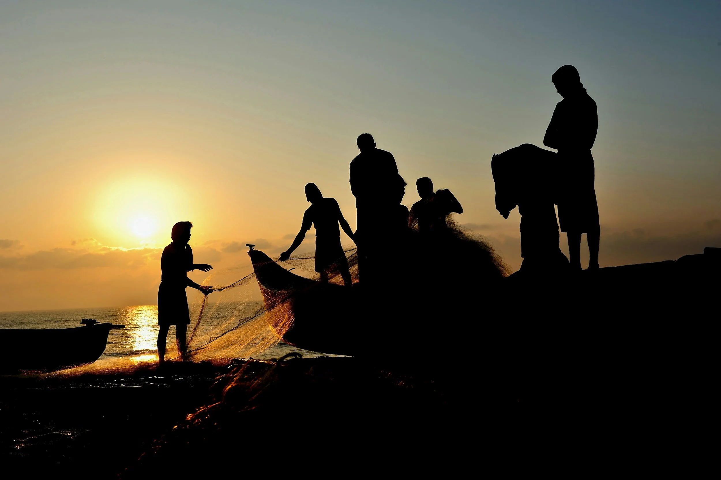 Silhouettes of people fishing and conversing on rocks at sunset by the ocean.