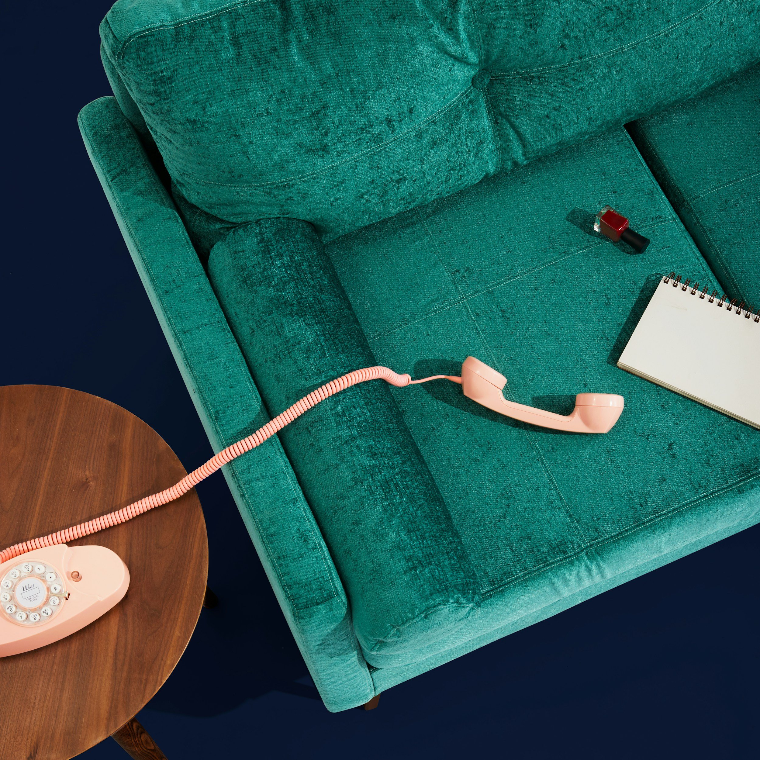 A pink rotary phone connected to a wooden side table with a coiled cord, placed on a teal sofa, with a lipstick and notebook nearby. EMDR. Anxiety. Shame. Women. NC. Therapy. Virtual.