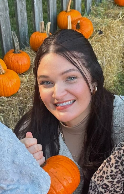 A smiling young woman with dark hair and blue eyes, sitting outdoors near a fence with pumpkins and hay, holding a pumpkin.