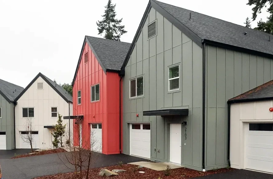 Row of modern townhouses with varying pastel-colored vertical siding, including white, red, and green, with small front yards, driveways, and attached garages.
