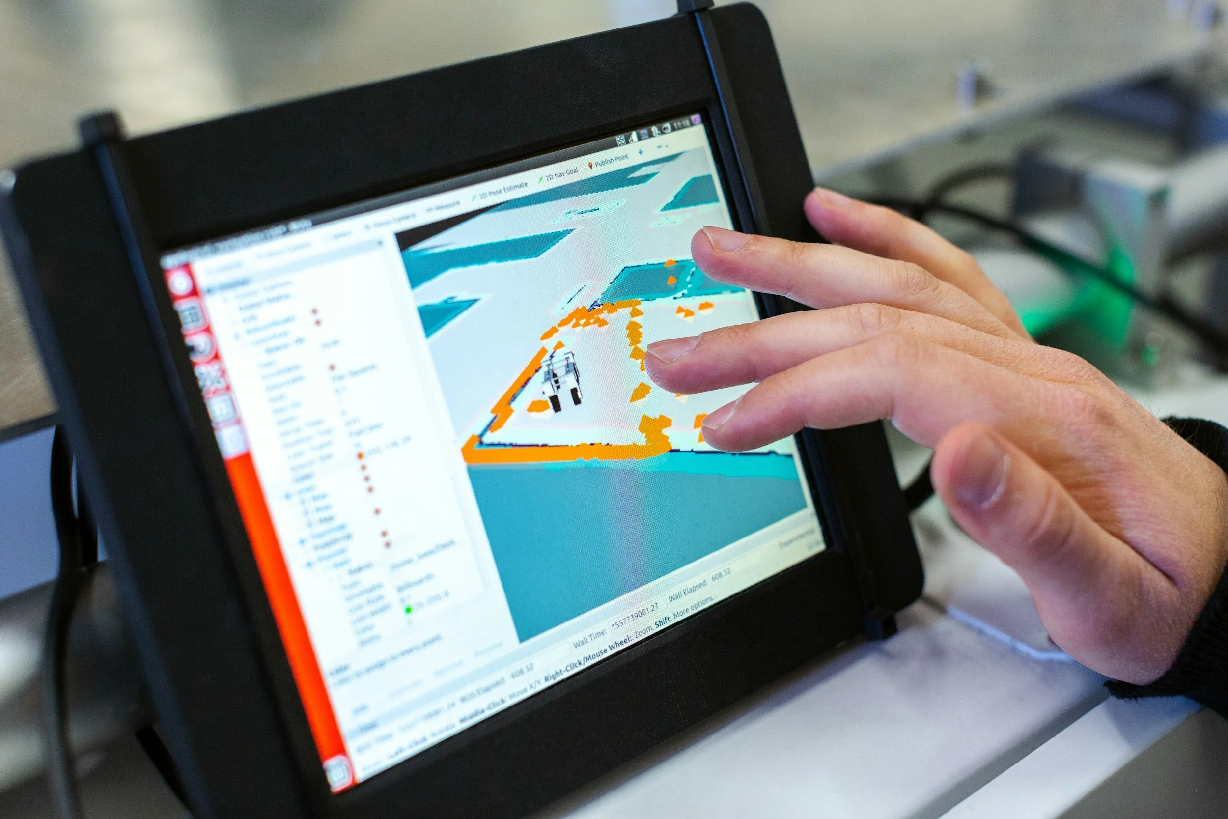 Person's hand using a tablet with a screen displaying a 3D mapping or simulation of a vehicle and environment.