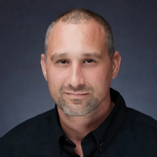 Headshot of a middle-aged man with short hair and a slight beard, wearing a black collared shirt, against a dark background.