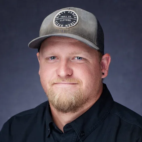 Man with a beard wearing a gray and black trucker cap and a black button-up shirt, against a dark background.
