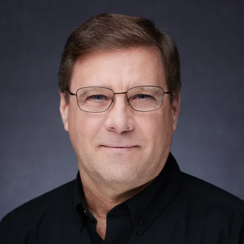 Headshot of a middle-aged man with brown hair, glasses, and a slight smile, wearing a black collared shirt against a dark background.