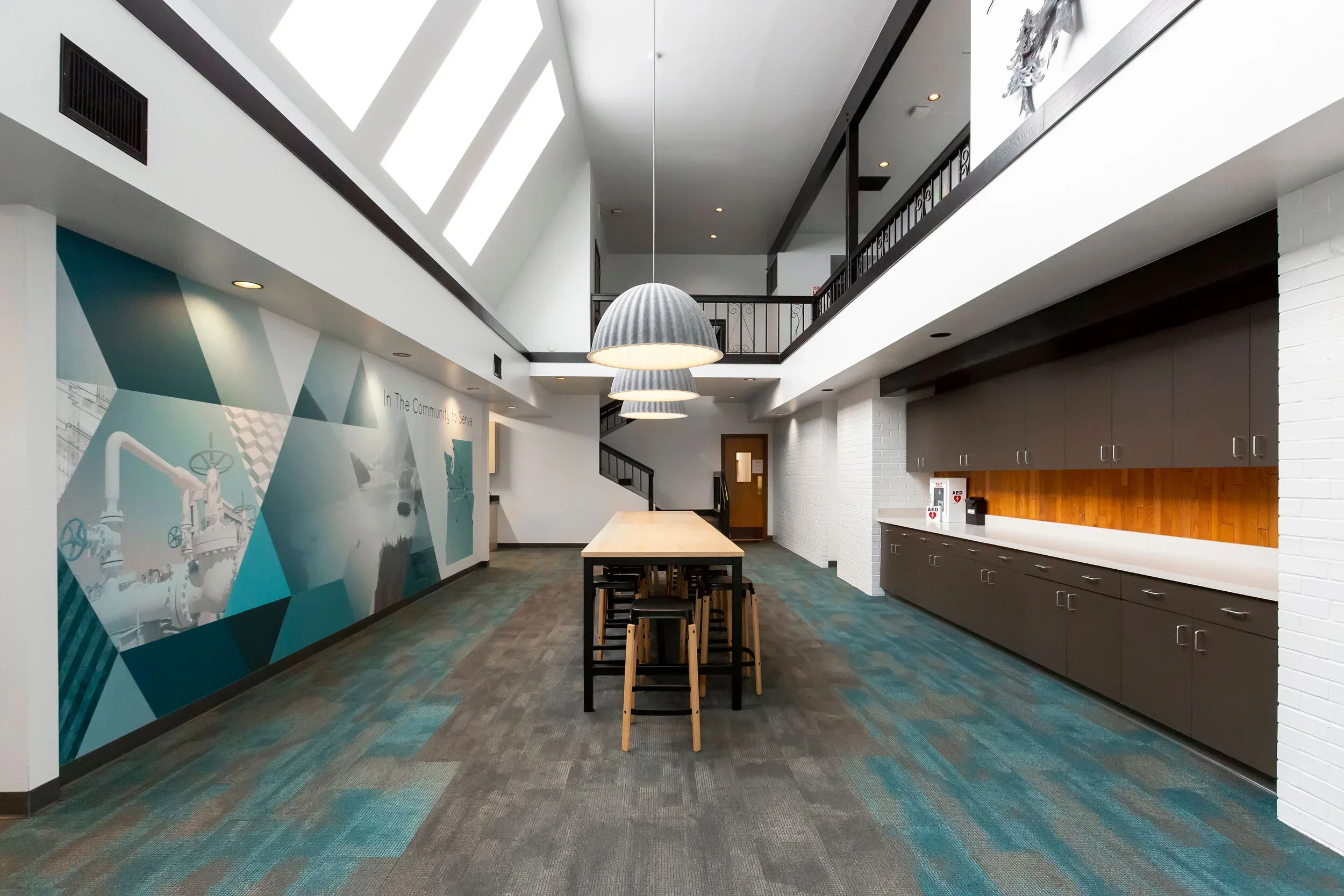 Interior of a modern, well-lit community space with a long table, hanging lamps, a mural on the wall, and a kitchenette area with cabinets.