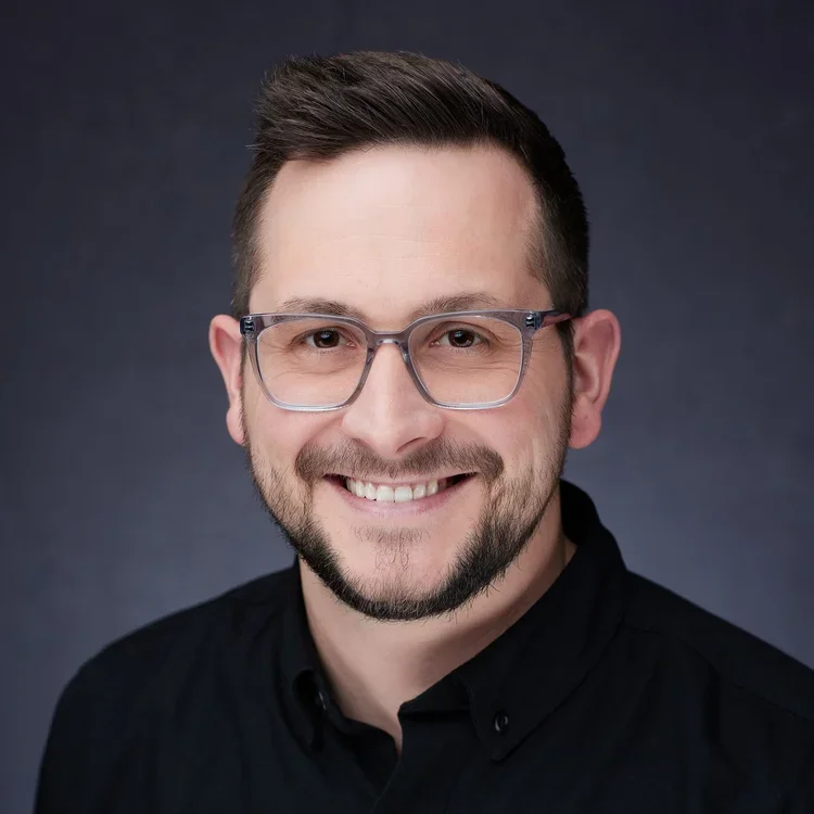 A smiling man with glasses, short dark hair, and a beard, wearing a black shirt, against a dark background.