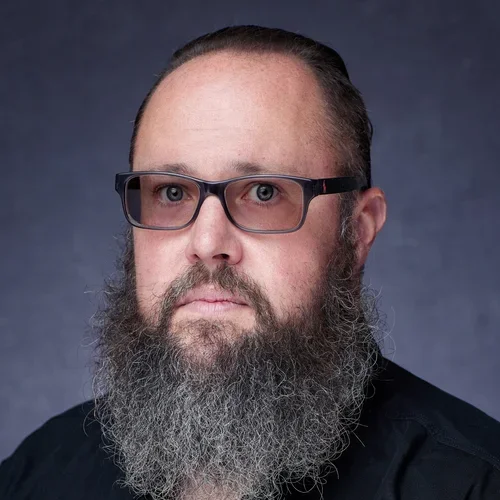 A man with glasses and a long gray beard posing for a portrait against a dark background.