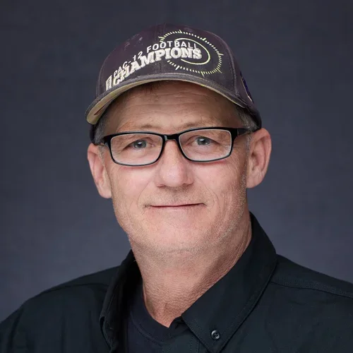A middle-aged man wearing glasses, a dark collared shirt, and a baseball cap that says "Super Bowl Champions" with a dark background.