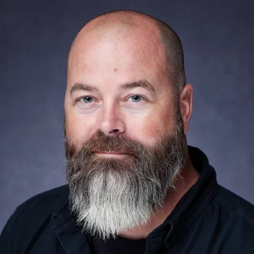 A middle-aged man with blue eyes, and a full beard, wearing a black shirt, against a dark background.